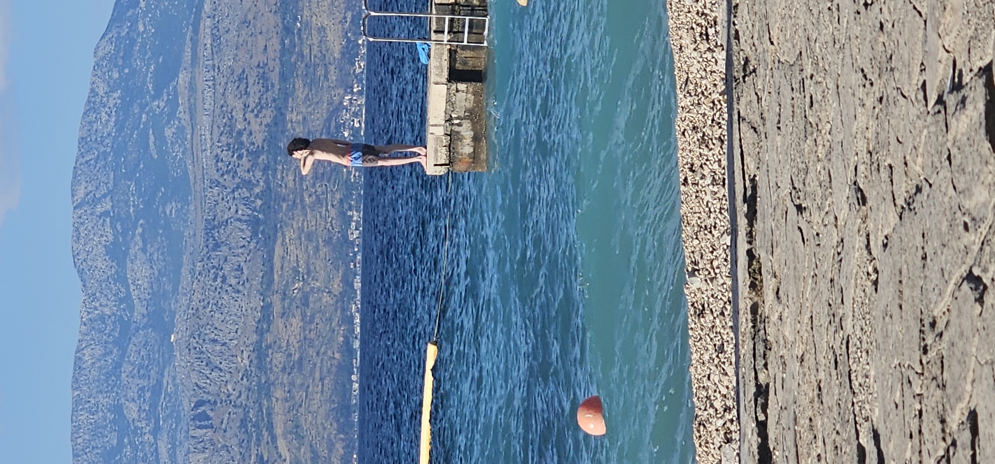 A young male, appearing to be of adolescent age, stands on a concrete pier overlooking a body of deep blue water. He is facing away from the camera, his right hand raised to shield his eyes from the sun as he looks towards the distant, rocky mountains and a coastal town. He is wearing blue and dark-colored swim trunks. The pier has a metal ladder extending into the water on its right side. A yellow floating barrier is visible in the water to the left of the pier, connected by a thin rope to a point further away. An orange buoy floats further out in the water. The foreground shows a shoreline covered in small, light-colored pebbles, bordering a textured concrete path. The background consists of the aforementioned mountains and the sky. The overall scene suggests a sunny day at a coastal location.