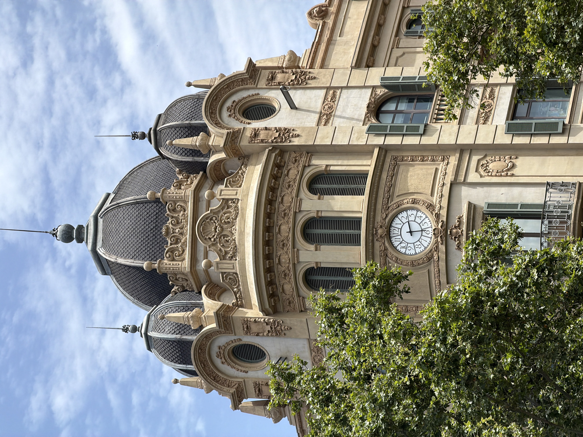 The image shows a close-up, low-angle view of the ornate facade of a building, with a focus on its architectural details. The building has a light-colored stone exterior and features multiple domed sections covered in dark, tiled scales. These domes are topped with spires. The facade is richly decorated with intricate carvings, including floral motifs, scrolls, and architectural moldings. Several windows with dark louvered shutters are visible, some with arched tops. A prominent feature is a large, round clock face with Roman numerals, situated on the building's facade. The clock hands indicate it is approximately 3:45. The scene is partially obscured by the lush green foliage of a large tree in the foreground, which casts shadows on the building. The sky above is a pale blue with scattered white clouds. This architectural style and the presence of lush trees suggest a Mediterranean setting, consistent with Barcelona, Spain. The image captures a moment during the daytime under fair weather conditions, highlighting the grandeur and historical character of the building. There is no visible text in the image.