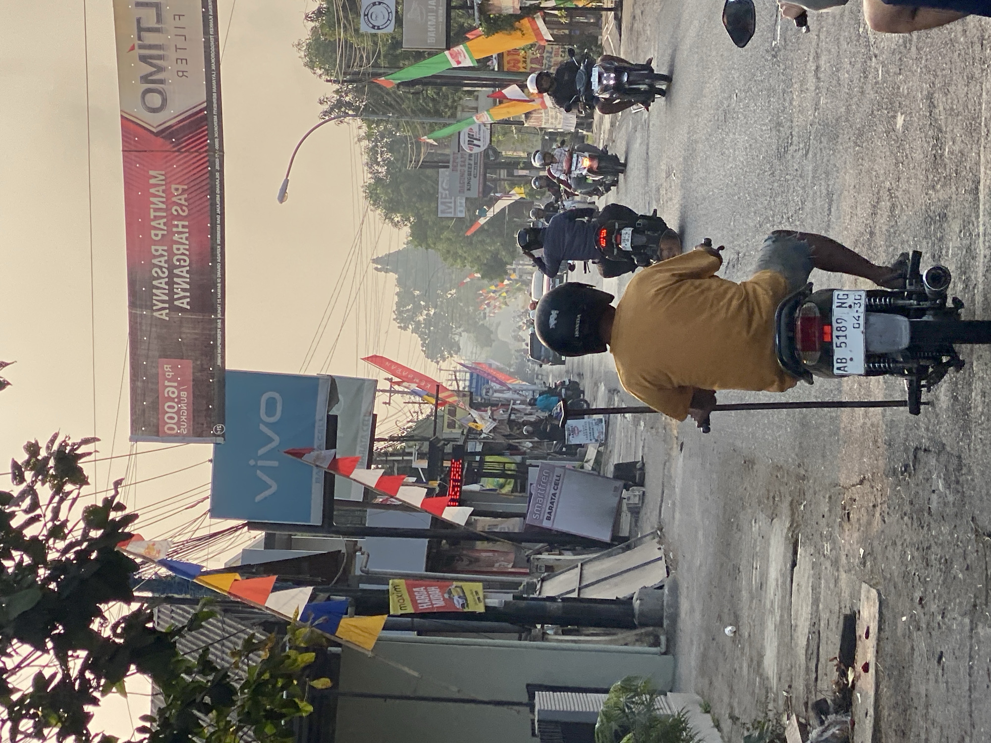 A street scene in Kasihan, Indonesia, captures a moment of daily life on a paved road lined with shops and decorated with numerous colorful flags. The time of day appears to be late afternoon, as indicated by the warm, soft lighting.
In the foreground, a person wearing a yellow t-shirt and denim shorts rides a dark-colored motorcycle, holding a long pole in their right hand, possibly for support or to indicate direction. Their back is to the camera, and their helmet is black. The motorcycle has a white license plate with the text "AB 5189 NG" and "04-3G" in black lettering.
The road is populated with several other motorcycles and riders. Some are moving in the same direction as the foreground motorcyclist, while others are coming from the opposite direction. The riders are wearing helmets, with varying styles and colors.
The buildings on either side of the street are mostly one or two stories high, with signs and advertisements displayed on their facades. Notable signs include a bright blue "vivo" logo, a "maxim HARGA MURAH" advertisement, and a "smartfren BARATA CELL" sign. There are also banners with Indonesian text, some of which appear to be advertisements or promotional materials.
The street is adorned with many small flags, primarily red and white, representing the Indonesian flag, as well as flags in yellow, blue, and orange. Electrical wires crisscross overhead, a common sight in urban Indonesian settings. The environment appears somewhat cluttered, with some debris visible on the side of the road. In the distance, more vehicles and buildings can be seen, suggesting a moderately busy urban environment. The overall impression is one of a lively, everyday street scene in an Indonesian city.