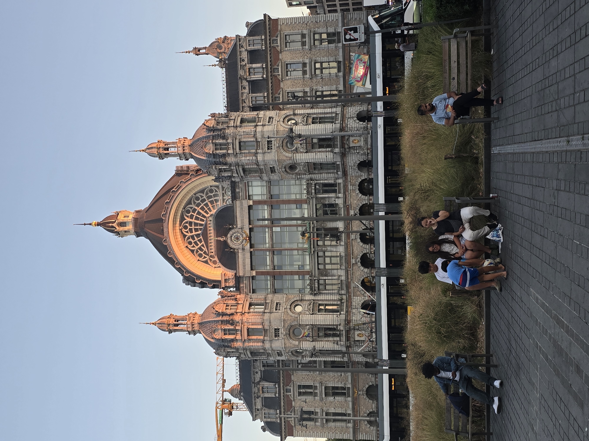 This is a medium shot, taken in the late afternoon, of the Antwerp Central Station in Antwerp, Belgium. The grand Beaux-Arts building dominates the background, its ornate facade bathed in the warm, golden light of the setting sun. The sky above is a clear, pale blue.
In the foreground, several people are seated on park benches. On the left, a young Black man with curly hair is sitting on a bench, looking at his phone, with a blue backpack beside him. To his right, a group of four people, appearing to be a family, are seated together. A young boy in a blue shirt and shorts is sitting on the lap of a woman, and they are playing with a soccer ball. Next to them, a young girl with glasses and a woman are also seated. On the far right, a man wearing a light blue polo shirt and dark pants is sitting on a bench, eating from a bag of chips. He is looking towards the right of the frame.
Between the people and the building, there is a strip of tall, dry grass. A modern tram is visible in front of the building, partially obscured by the grass. A construction crane is visible to the left of the station building. The ground in the foreground is paved with dark grey bricks.