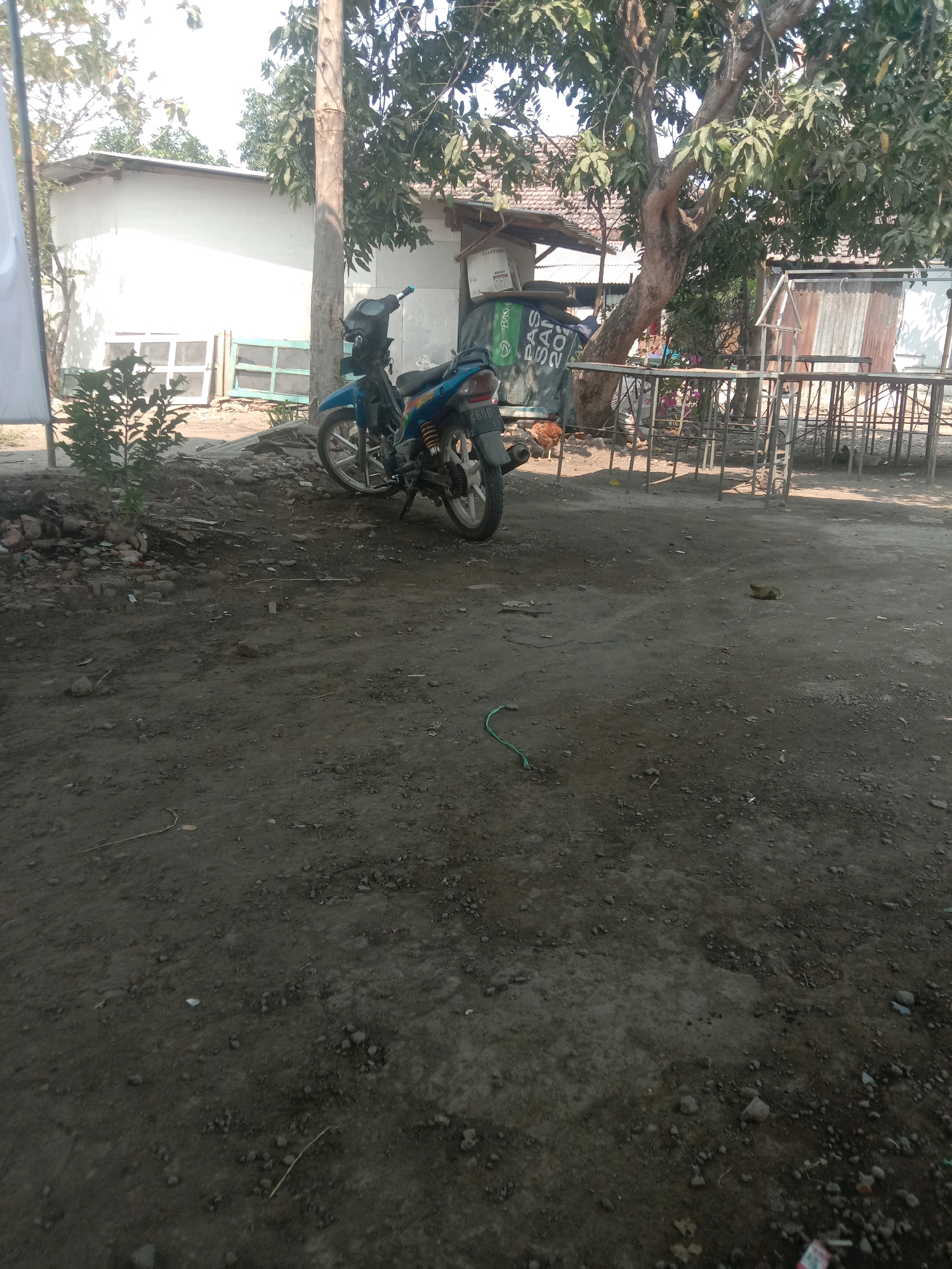 A motorbike is parked on a dirt road in Karanganyar, Indonesia. The motorbike is blue and black and is facing to the left. Behind the motorbike, there is a white building with a white roof and a large tree. There are also some metal structures and a fence in the background. The ground is covered in dirt and small stones. There are a few chickens visible in the background. The sky is clear and the sun is shining.
