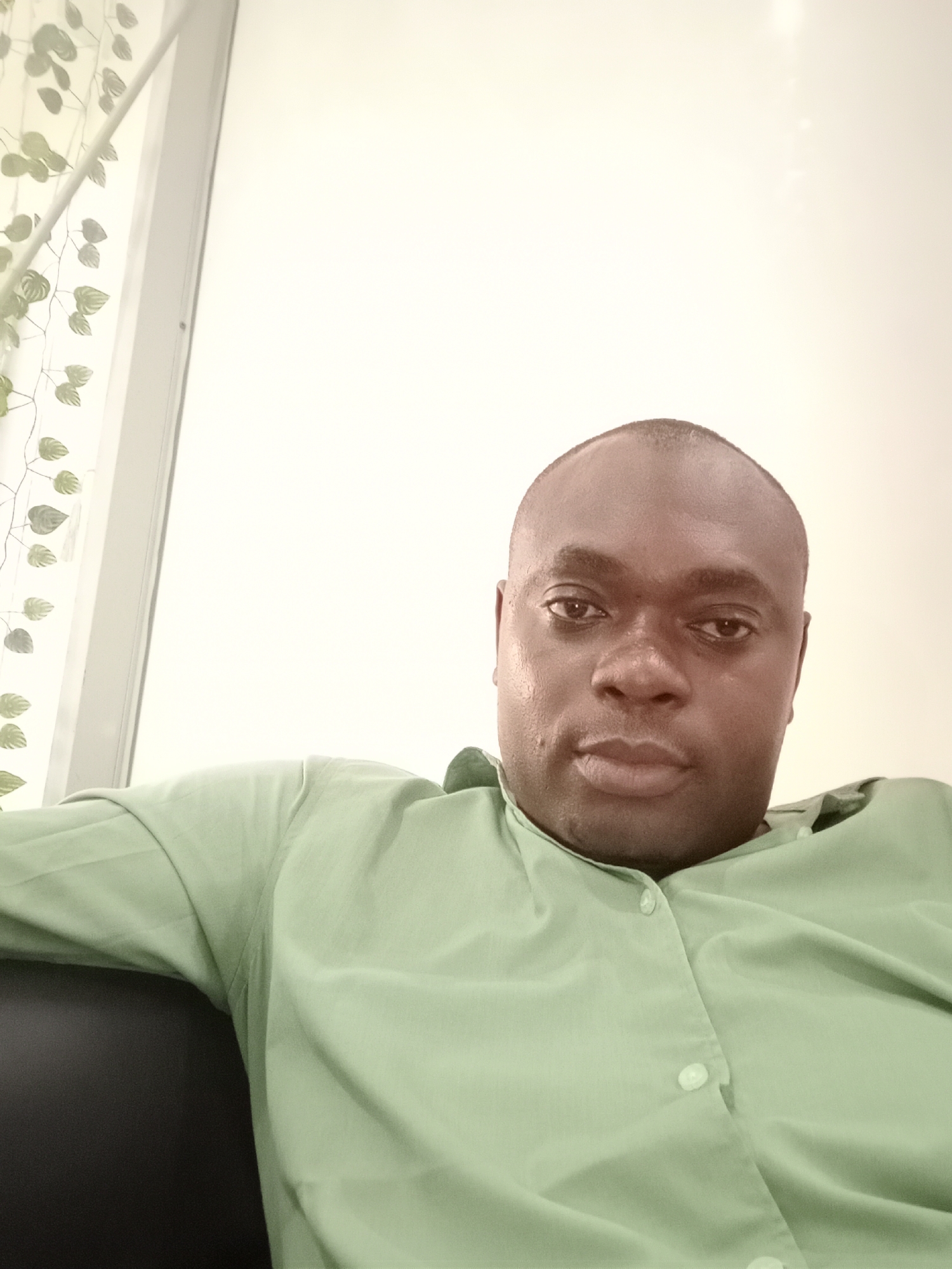A medium shot depicts a dark-skinned man wearing a light green button-down shirt, seated indoors. The background features a white wall adjacent to a window, which has a leafy design on the glass or covering it. The location is not apparent from the image.