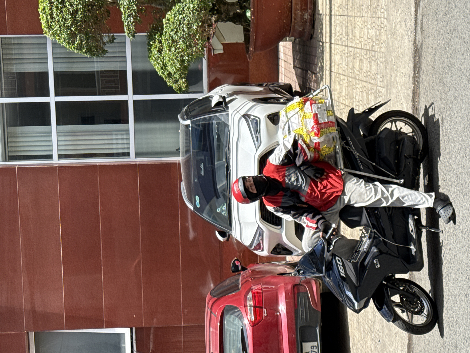 A person wearing a red and black jacket, white pants, a red helmet, and a black face mask sits on a black motorcycle. The motorcycle is parked on a sidewalk next to a red car and a white SUV. A large potted tree is visible to the right of the SUV. The background features a building with a red brick facade and large windows with vertical blinds. The scene appears to be in an urban setting in Da Lat, Vietnam, likely a street or parking area.