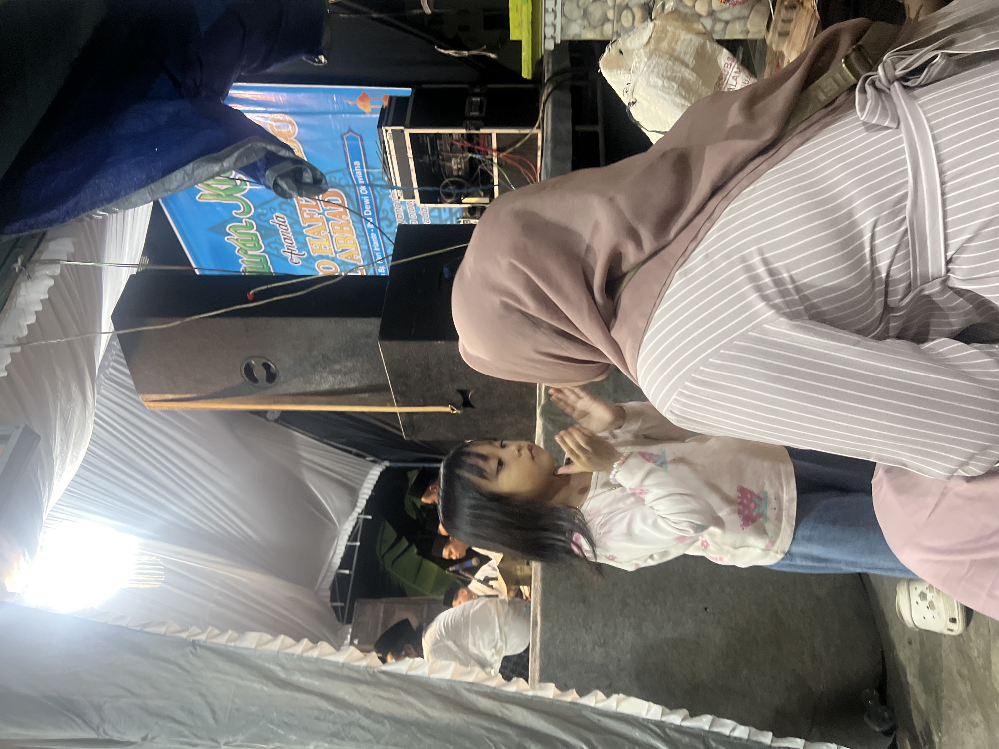 In Wedarijaksa, Indonesia, a young girl with dark hair stands clapping her hands in front of an adult woman wearing a light brown hijab and a striped dress. The girl is wearing a light-colored shirt with a floral pattern and blue pants. Behind them, a large speaker system is visible, along with a blue banner featuring Arabic script and text that appears to be an announcement. The scene is set outdoors under a temporary tent or canopy, with white fabric draped overhead. The ground appears to be concrete. Several other people, some wearing peci hats, are visible in the background, possibly participating in or observing an event. A white clog is lying on the ground near the girl's feet.