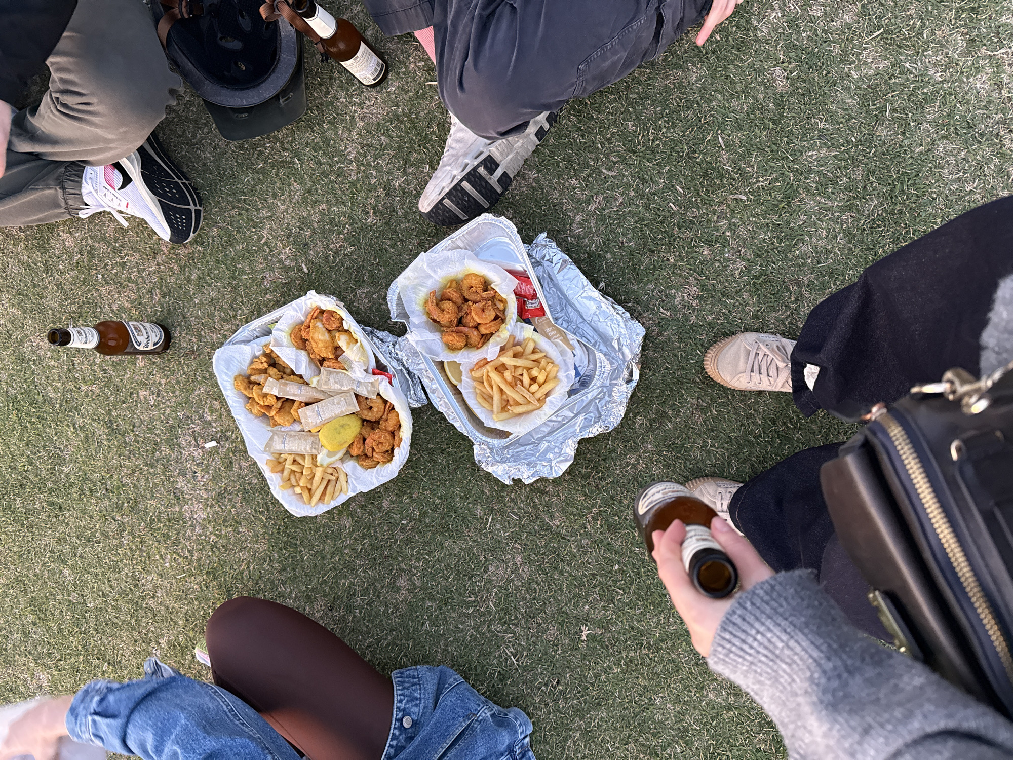The scene depicts an informal gathering of individuals on a grassy surface, likely a park or lawn, though the specific location in Tel-Aviv, Israel, is unidentifiable from the image. Several people are seated or positioned around two aluminum foil-lined trays of food, containing fried shrimp, fried fish, and french fries. Condiment packets are also visible in the trays. Empty and full beer bottles are scattered around. The individuals are only partially visible in the frame, showing details like shoes, clothing, and hands holding a beer bottle. A black bag or purse is also visible.