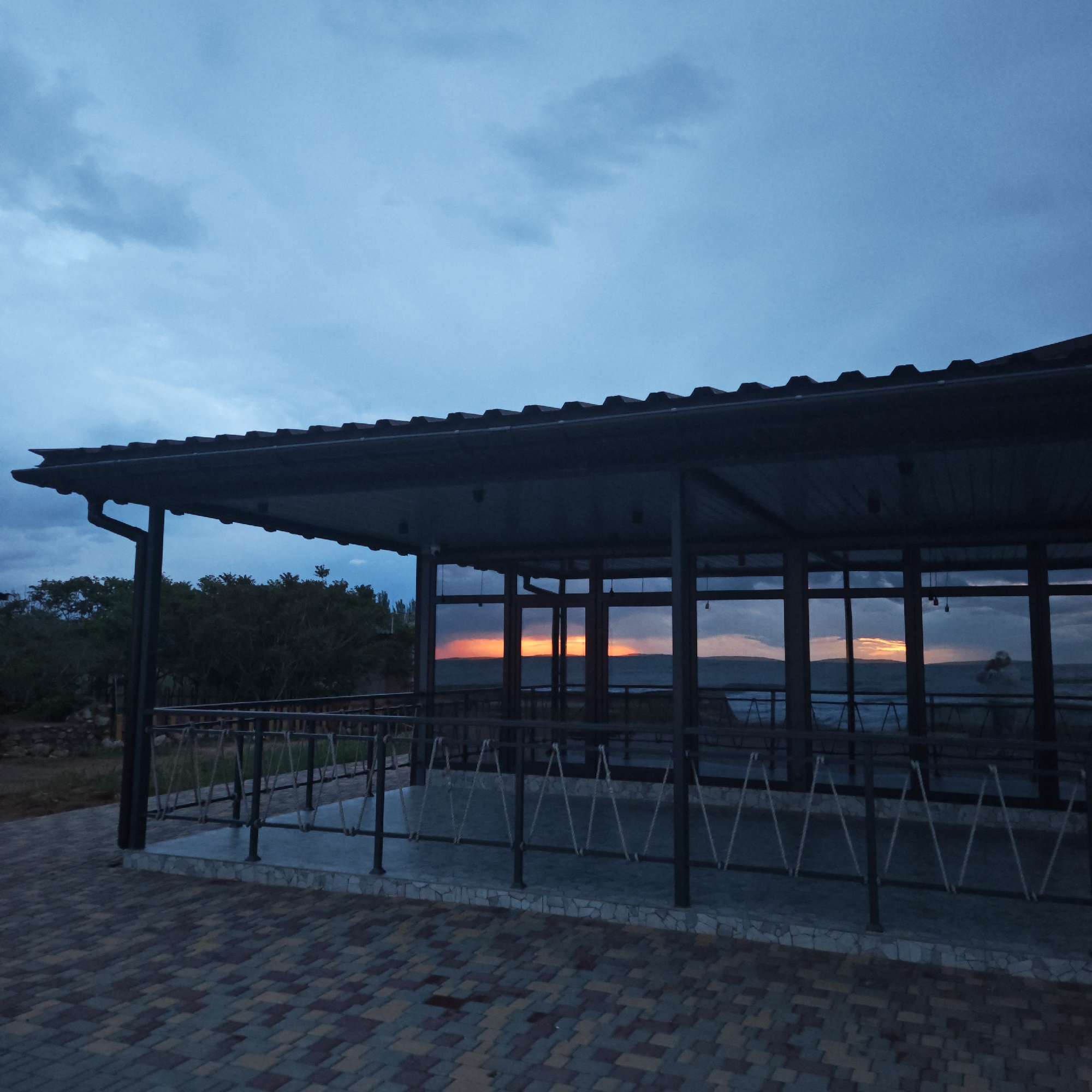 The image depicts an outdoor pavilion in городское поселение Щелкино, Ukraine, at twilight. The pavilion has a dark metal roof and frame, with large glass windows that reflect the sunset over the water. The sunset is visible through the windows as a horizontal band of orange and red light. A metal railing with rope accents surrounds the elevated platform of the pavilion. The ground is paved with patterned bricks in shades of brown, gray, and tan. In the background, trees are silhouetted against the sky. The sky is a gradient of blues and grays with heavy cloud cover.