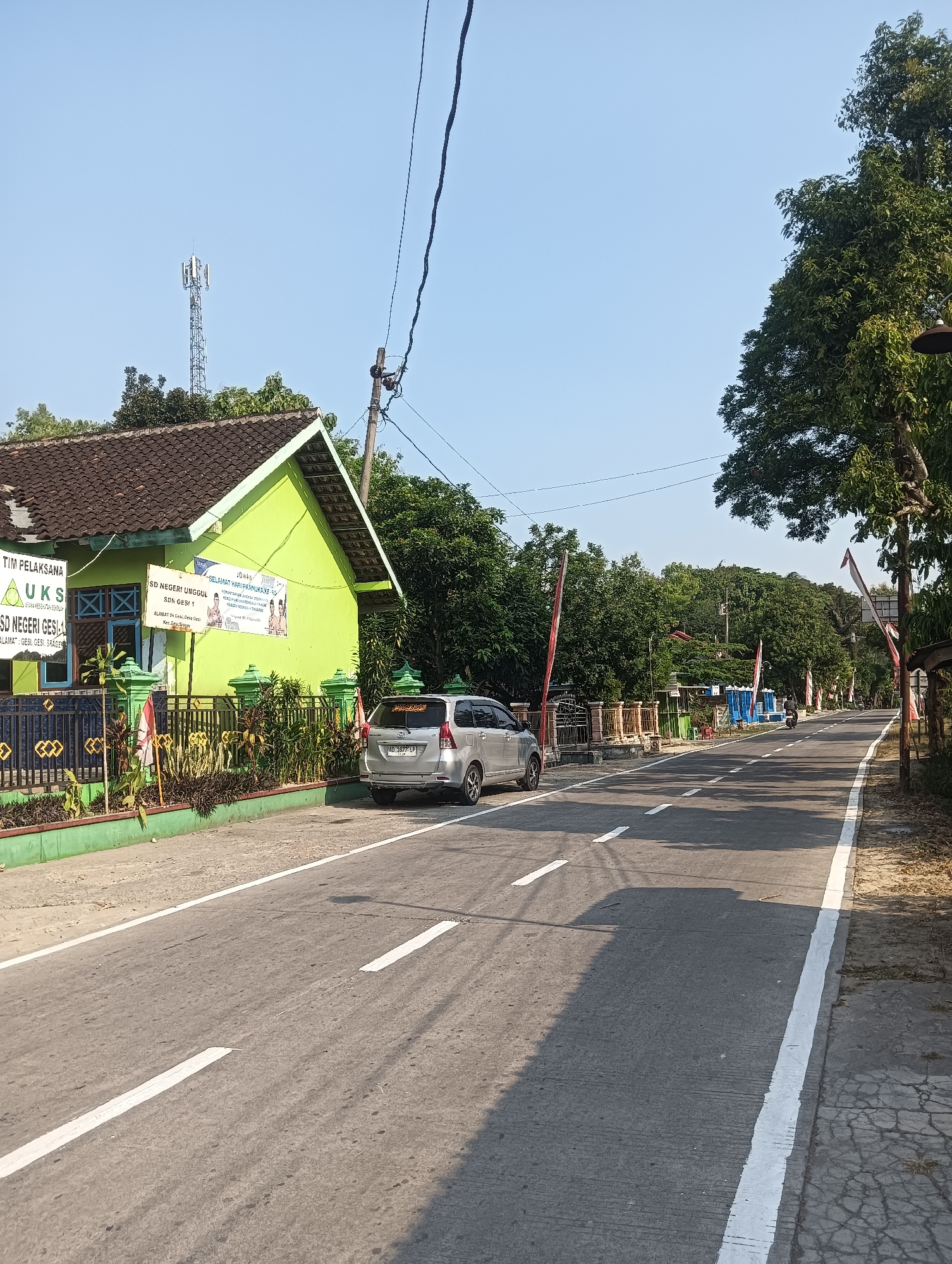 The image captures a street scene in Palemrejo, Indonesia, on a clear, sunny day. On the left side of the road, a bright green building, identified by signage as "SD NEGERI UNGGUL" (which translates to "Superior Public Elementary School"), stands with a dark tiled roof. In front of the school, a black metal fence encloses a garden area with various plants and a few Indonesian flags. A silver minivan is parked to the right of the school entrance. The road itself is asphalt, marked with white lane dividers. Further down the road, trees line both sides, and a few small structures and shops are visible. A telecommunication tower can be seen in the distance against the light blue sky. A single motorcyclist is present on the road, heading away from the viewer. The overall impression is a quiet, rural or suburban street on a bright morning.