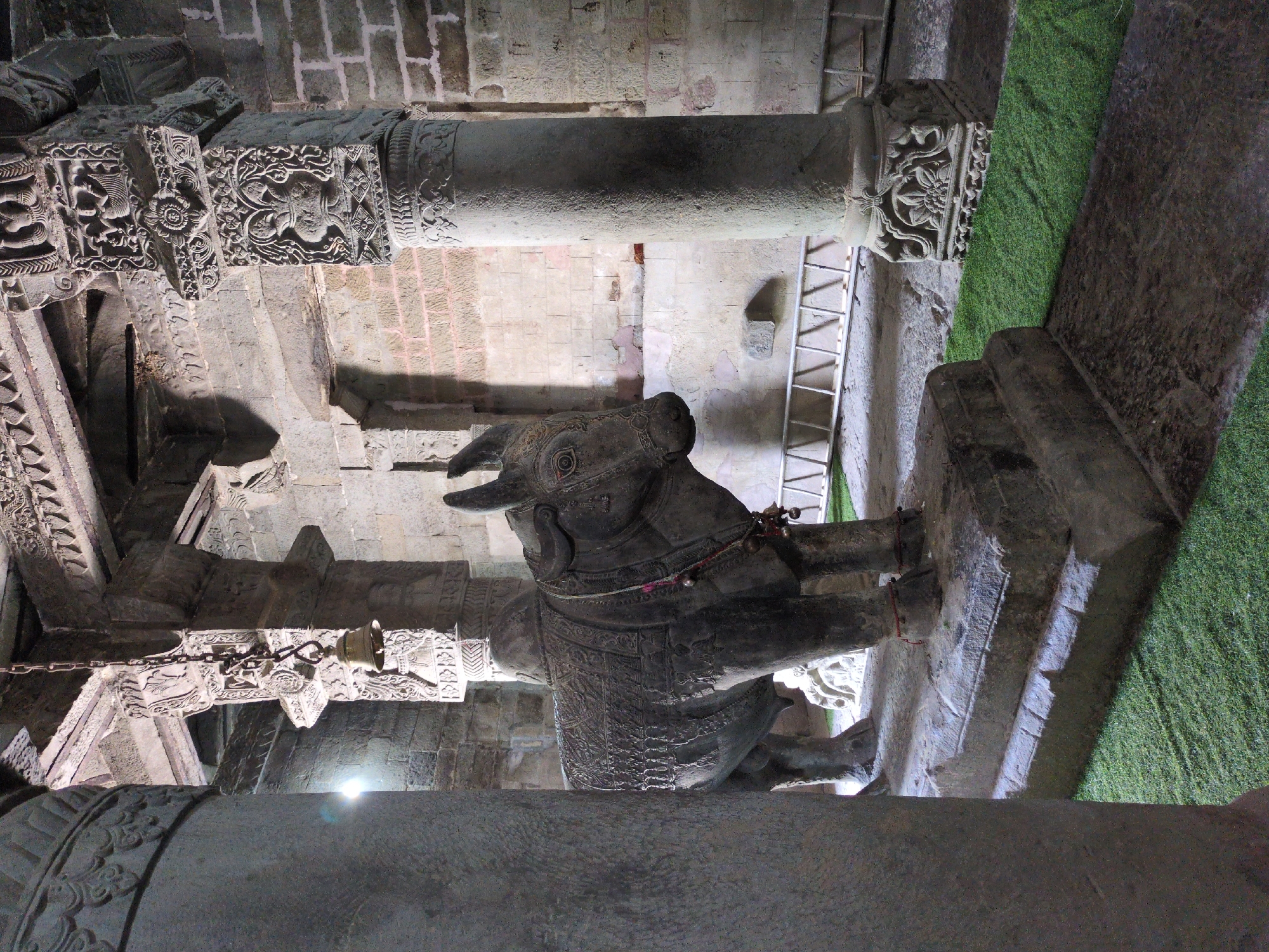 A stone sculpture of a bull, adorned with a decorative saddle cloth and bells, stands on a stone platform in an interior space. The bull is positioned in the center of the frame, facing right. The structure features large stone pillars with intricate carvings, a stone ceiling with decorative elements, and brick walls in the background. Green artificial turf is laid out on the floor in front of the bull and around the pillars. A chain with a bell hangs from the ceiling above and slightly to the left of the bull. A metal ladder is visible in the background, leaning against a wall. The lighting appears to be artificial, creating shadows and highlighting the textures of the stone. The overall scene suggests a temple or a historical site.