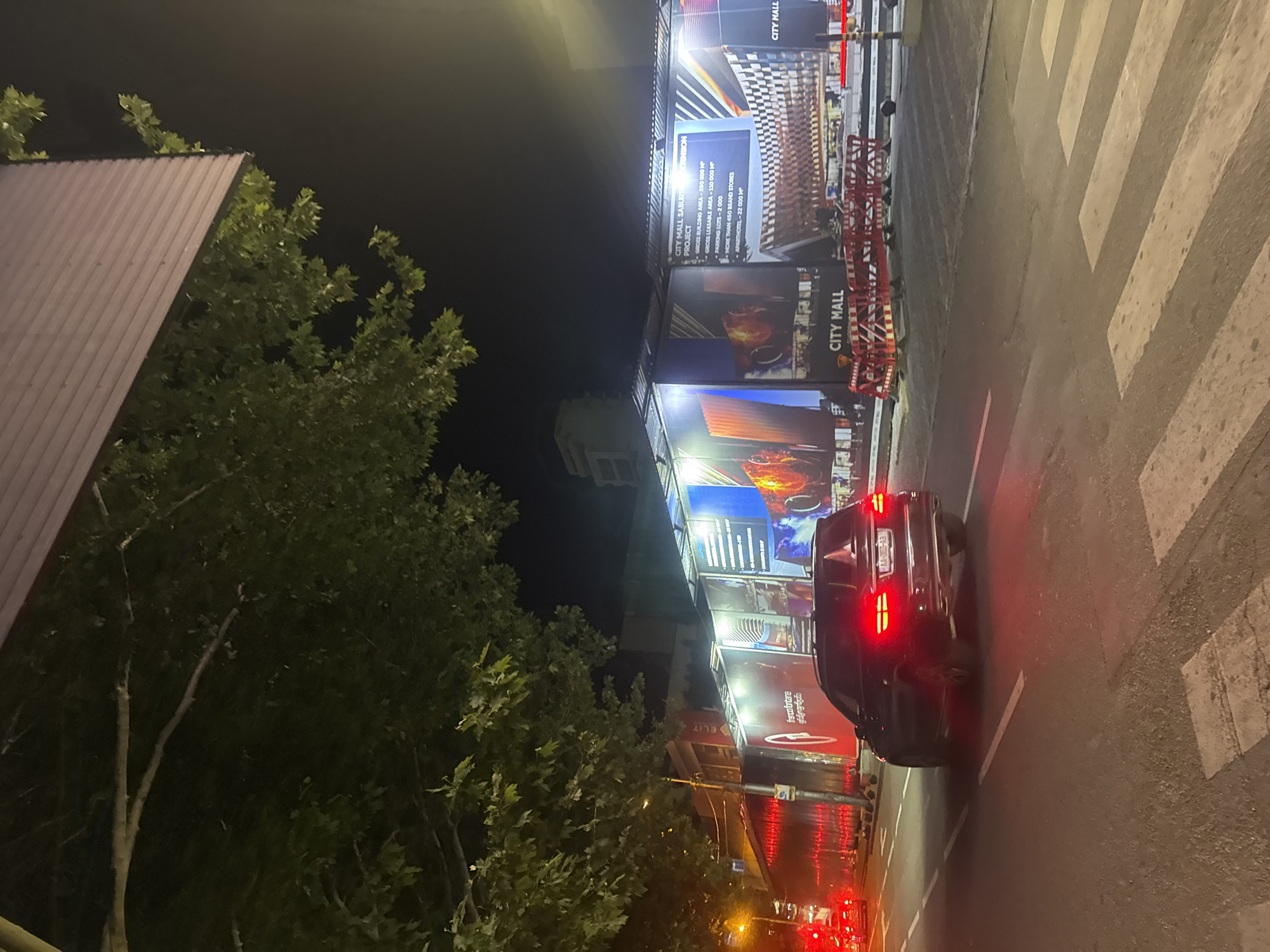The image depicts a night scene on a street in Tbilisi, Georgia, likely near a construction site for the "City Mall Saburtalo Extension." A dark-colored SUV, possibly a Mercedes-Benz based on its taillight design, is driving away from the camera, its brake lights illuminated. The vehicle has a license plate that reads "AA-003-AA." The street features a pedestrian crossing with white stripes. To the right of the road, a large construction barrier with bright red and white warning signs is visible, suggesting ongoing work. Behind the barrier, large printed banners display architectural renderings of the "City Mall Saburtalo Extension" project, detailing specifications like gross building area, leasable area, parking capacity, and the number of brand stores and apartments. Trees with green foliage overhang the scene from the left, and some building structures are visible in the background, illuminated by streetlights and the construction site's lighting. The overall ambiance is urban and suggests activity despite the late hour.