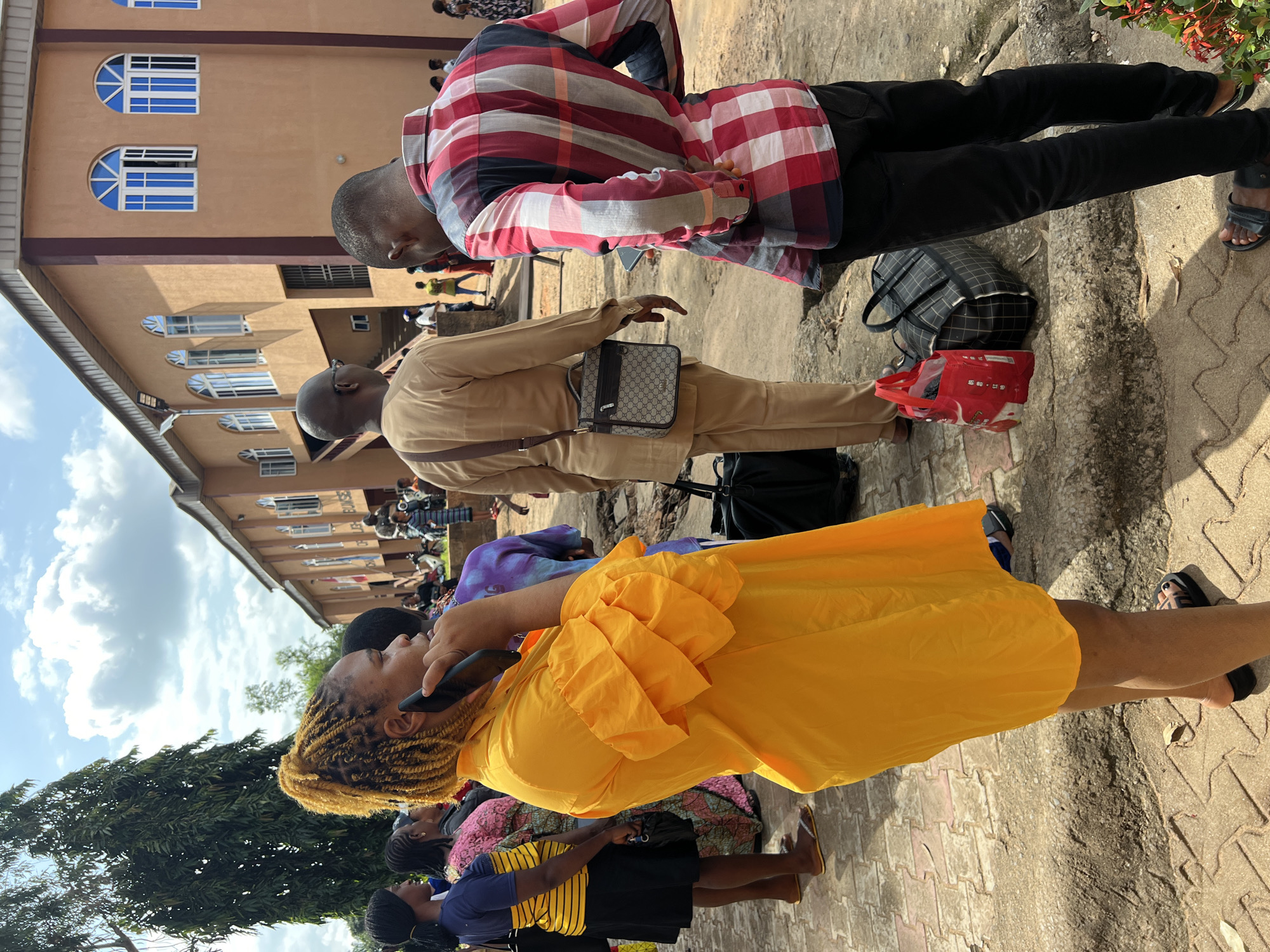 In Nnewi, Nigeria, a group of people are gathered outdoors in front of a building with a light brown facade and arched windows. A woman in the foreground, wearing a bright yellow dress with ruffled sleeves, is on a phone call. She has blonde braided hair and is talking into a dark-colored smartphone. To her right, a man in a tan traditional outfit with a crossbody bag stands with his back to the camera. Next to him, a man in a red, white, and black plaid shirt and black pants is also standing, facing away. Several large bags, including a checkered duffel bag and a red shopping bag, are placed on the ground near the two men. Other individuals, some dressed in vibrant clothing, are visible in the background. The ground is paved with interlocking bricks and stones, and there is vegetation and a large tree to the left. The sky above is partly cloudy.