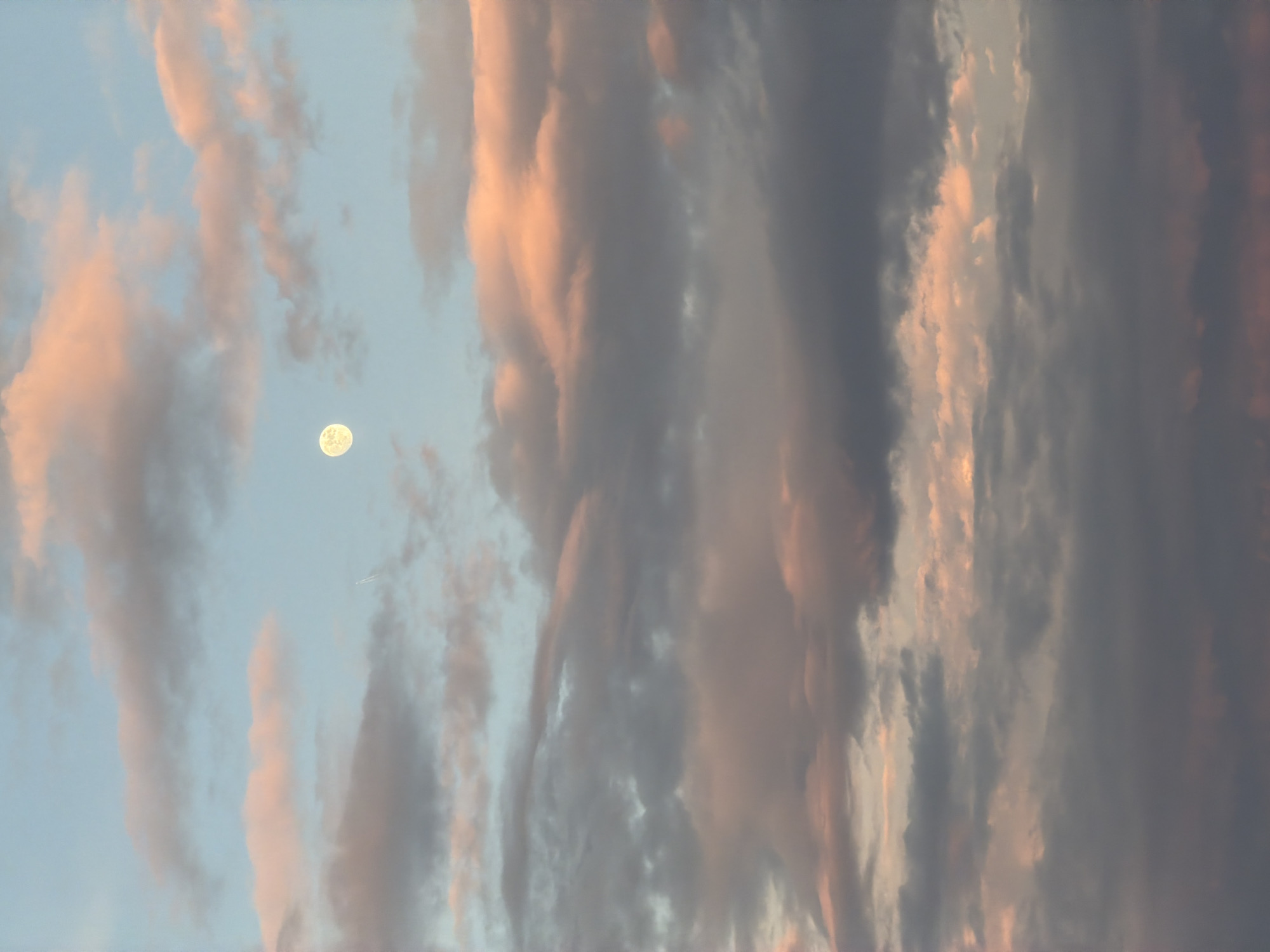 The image depicts a vertical view of a sky filled with layered, striated clouds in shades of grey, pink, and peach, indicative of either sunrise or sunset. A full moon is visible in the left-center area, set against a patch of pale blue sky. A distant contrail, likely from an airplane, is also visible. The lack of any ground-level elements makes it impossible to confirm the location in the city of Budva, Montenegro.