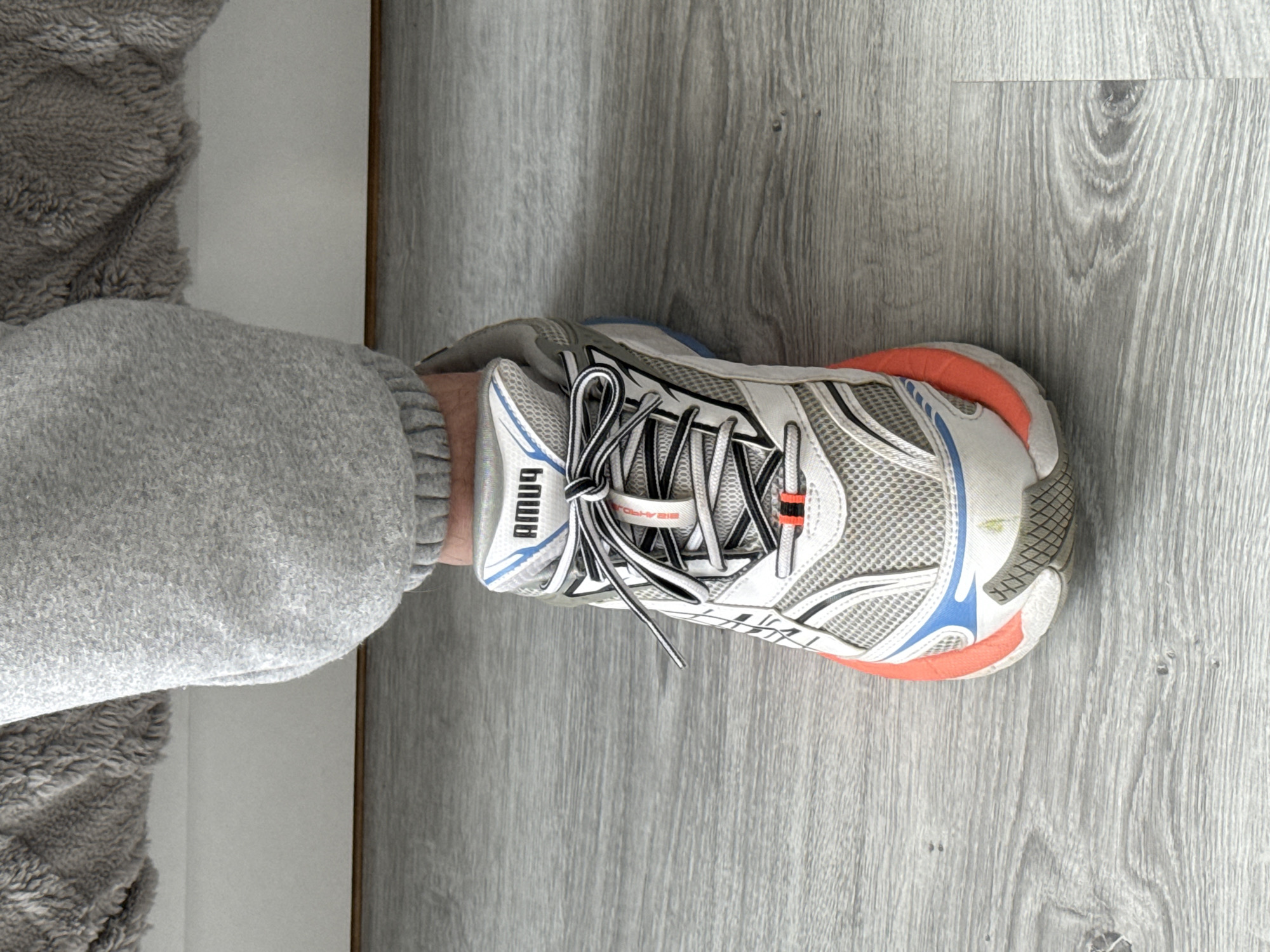A photograph depicts a person's leg, clad in grey sweatpants, with a white and grey running shoe on the foot. The shoe has black and white striped laces and orange and blue detailing. The word "AMU4" is printed vertically on the side of the shoe near the ankle. The shoe is positioned on a grey, wood-like surface. The edge of a white surface is visible next to the sweatpants. There is no indication that this image was taken in Stara Zagora, Bulgaria. The objects are, in order of visual prominence, a shoe, a leg with a sweatpant, and a floor.