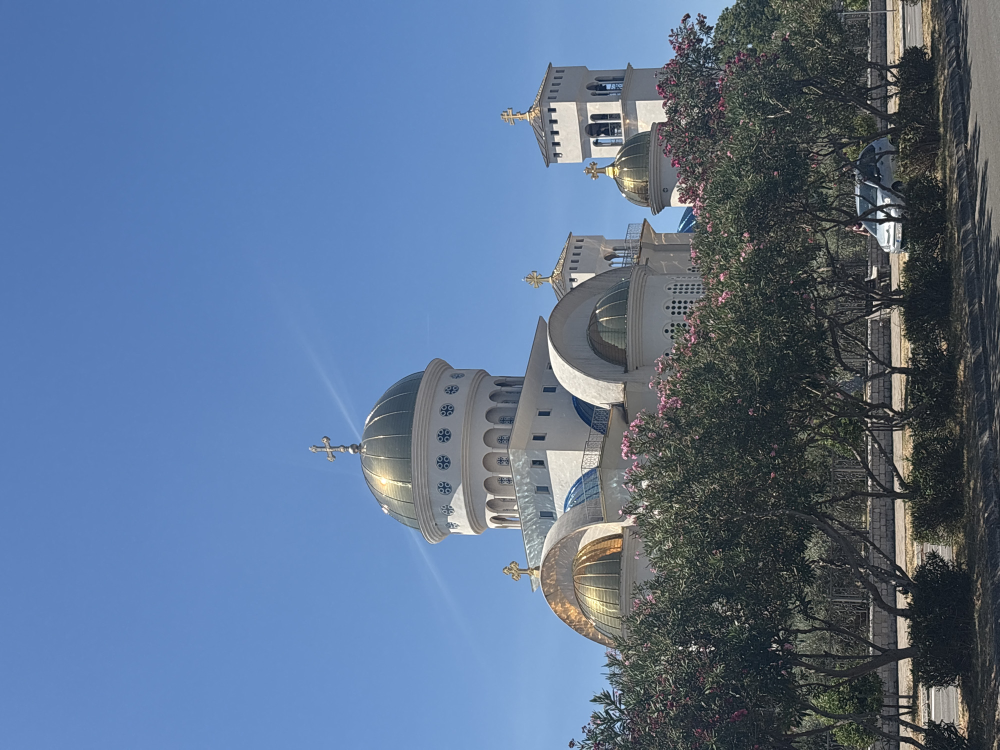 A bright, sunny day in Bar, Montenegro, features the imposing presence of the Cathedral of the Holy Trinity. The large, white, multi-domed church is adorned with prominent gold-colored domes topped with crosses, reflecting the clear blue sky. Lush green trees and shrubs, some with delicate pink flowers, form a verdant foreground, partially obscuring the lower levels of the church and a stone wall behind them. A paved road runs in front of the vegetation. In the middle distance, a silver car is parked. The overall scene captures a serene, architectural landmark set against a vibrant natural backdrop.