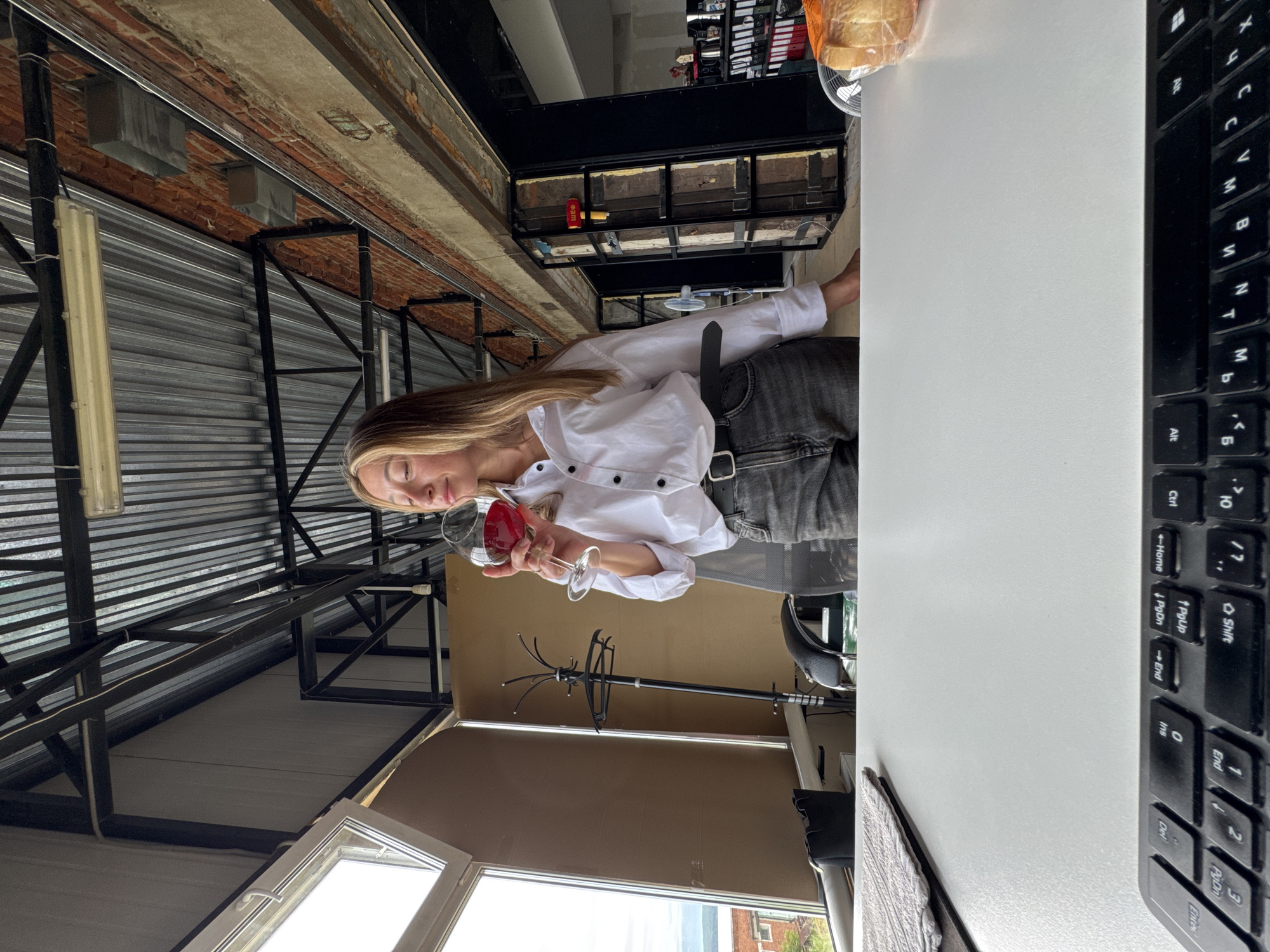 A woman with blonde hair, wearing a white collared shirt and dark jeans, stands in an industrial-style loft space. She is holding a wine glass filled with red wine, with her eyes closed and a slight smile on her face. The background features exposed brick walls, metal beams, a corrugated metal ceiling, and large windows. A coat rack, a desk with a keyboard, and shelves stocked with binders are visible. A loaf of bread in plastic wrap sits on the desk to the right. The scene appears to be an office or workspace, possibly with a view of the city of Chaikovsky, Russia, visible through the windows.