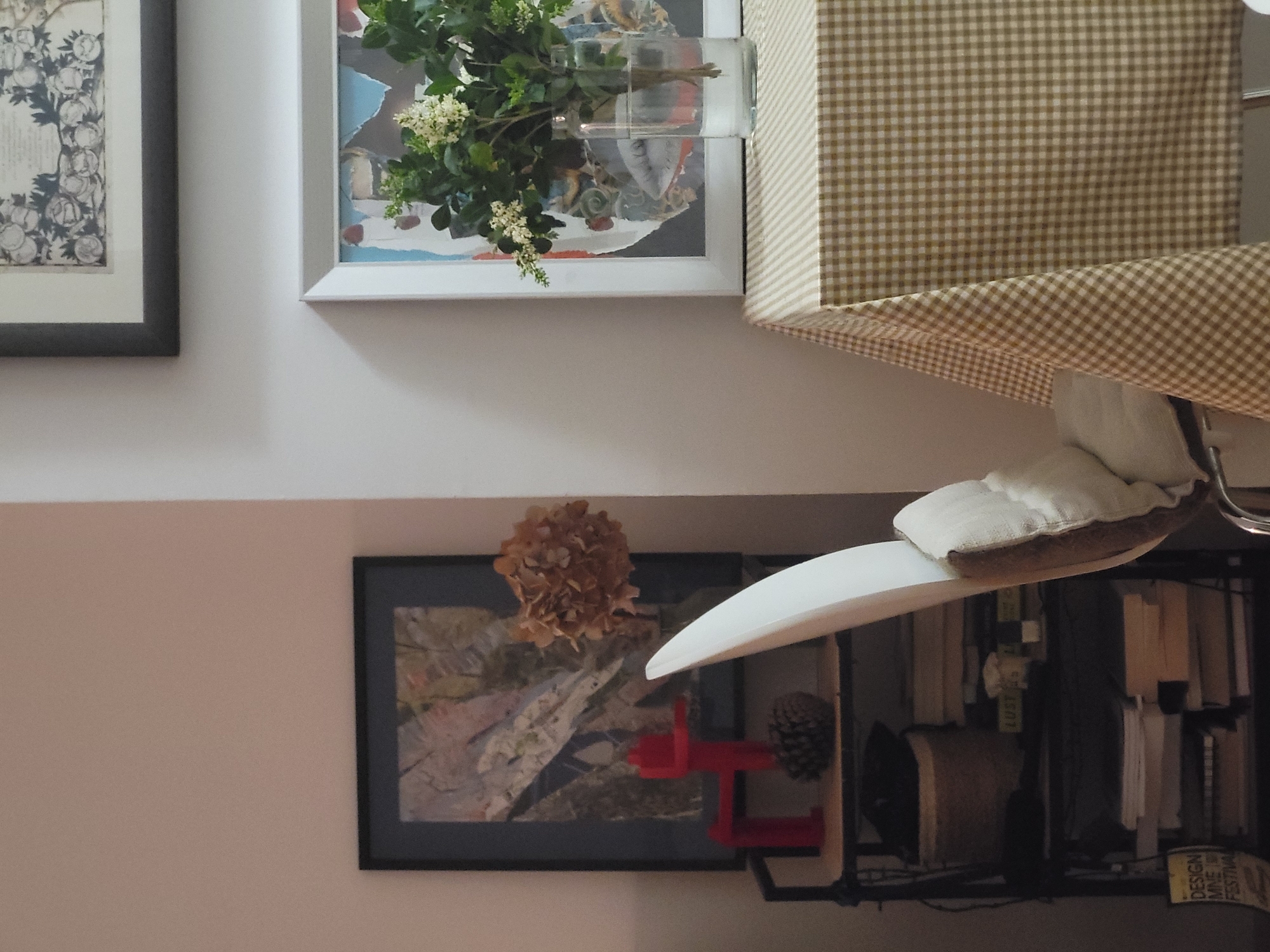 The image shows a portion of a wall with various objects hanging on it. On the upper left, there is a framed print in a dark frame. To the right of this, another framed picture shows a vase with flowers. A brown and white gingham patterned curtain or valance is partially visible on the upper right.
Below the prints and curtain, there is a dark-framed picture. Above it is a dried flower. Next to the picture is the white seat of a chair, and a black shelf holding books and other objects. A red toy is visible next to a pine cone, sitting on the shelf. The writing "DESIGN MNE 14/20 FESTIVAL" is visible in yellow on the lower right.
The image is taken indoors, and there is no information to determine if the location is in Budva, Montenegro.