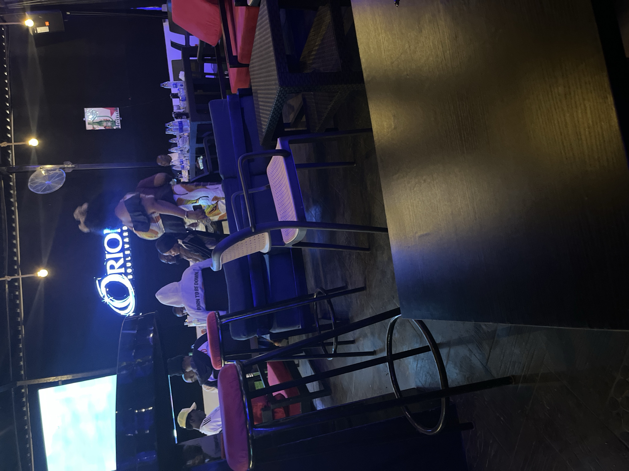 This is a mid-shot, eye-level, outdoor evening scene at Orion Boulevard, a nightlife establishment in Abuja, Nigeria. The focus is on the seating area and a few patrons. In the foreground, two red-topped bar stools with black metal frames are prominently featured. To the right, a black woven-style coffee table sits between two red armchairs. Further back, a group of people are seated at a dark-colored table laden with water bottles. A woman in a white hoodie with "BORN TO BE DOWN" printed on the back is visible. In the background, a large screen displays a bright blue and white graphic, presumably the Orion Boulevard logo. A ceiling fan is mounted above and to the left of the screen. A poster for Heineken is affixed to the dark wall on the right side of the image. The overall ambiance is dimly lit, with various light sources contributing to the mood, including ceiling lights and the glow from the screen. The ground appears to be tiled.