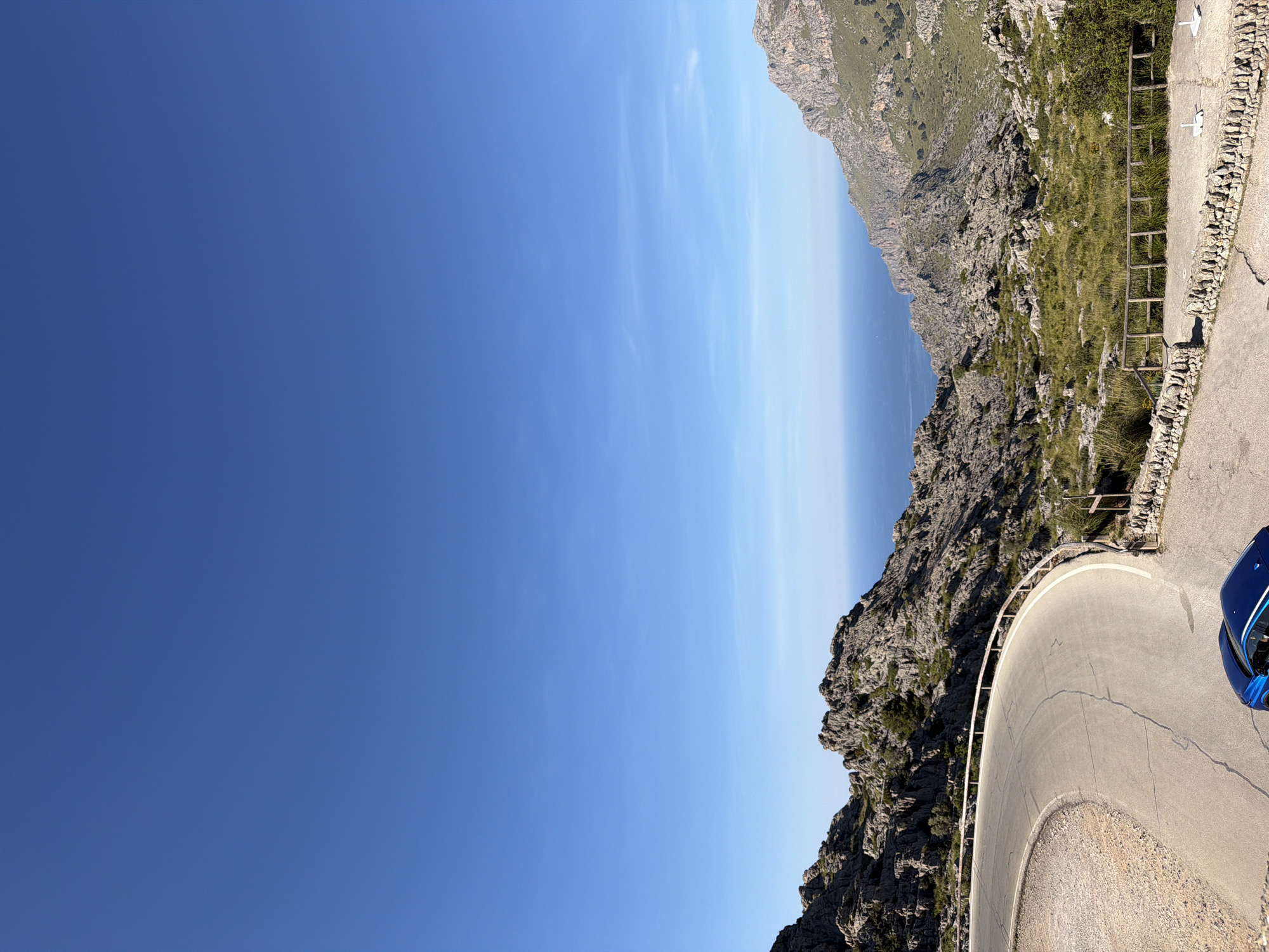 The image shows a scenic view of a winding road in the city of Escorca, Spain. The road is paved and curves around the mountainous terrain. On one side of the road, there's a steep, rocky cliff covered with sparse vegetation. Beyond the cliff, a glimpse of the sea is visible, creating a horizon line. The sky is clear and blue, suggesting a sunny day. A portion of a blue car is visible on the side of the road. A wooden fence borders the road edge in some areas.