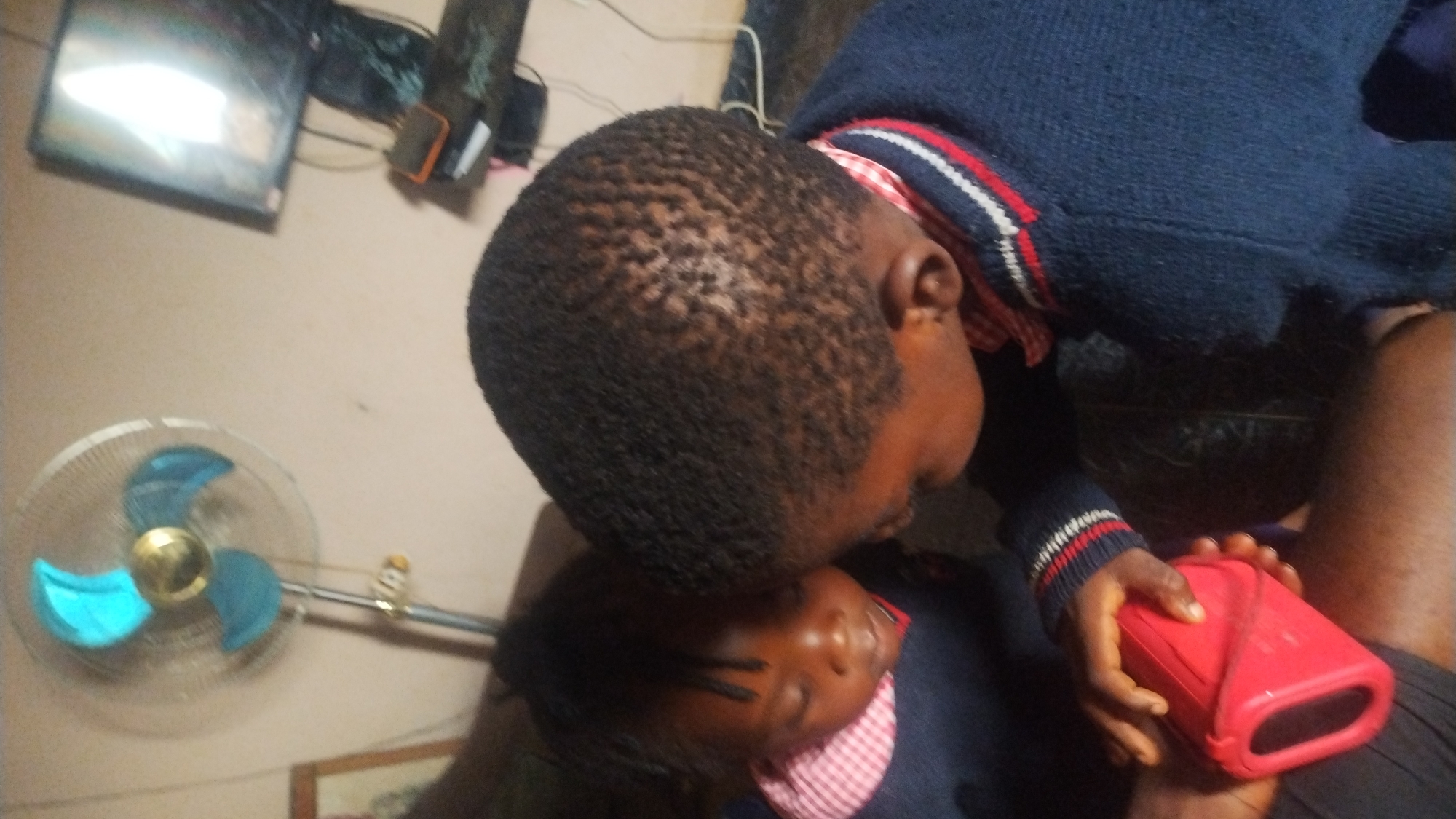 The image is rotated 90 degrees counter-clockwise, depicting an intimate indoor scene with two individuals. One person, positioned towards the right of the frame, wears a dark blue knitted sweater with red, white, and blue striped cuffs, and a red and white checkered garment underneath. This individual has short, dark, tightly curled hair and holds a red portable speaker in their right hand. Their head is bowed, looking down.
The second individual, partially visible beneath the first, has dark hair and is also wearing a red and white checkered garment. Their eyes appear closed or are directed downwards, resting against the first person. The pose suggests comfort, closeness, or a moment of rest.
In the background, on the left, a floor fan with blue blades and a gold center is visible. Above the fan, mounted on the wall, is a television screen reflecting a bright light. Cables and other objects are clustered near the television. The setting appears to be an indoor environment, likely a home. No discernible text is visible on any objects.