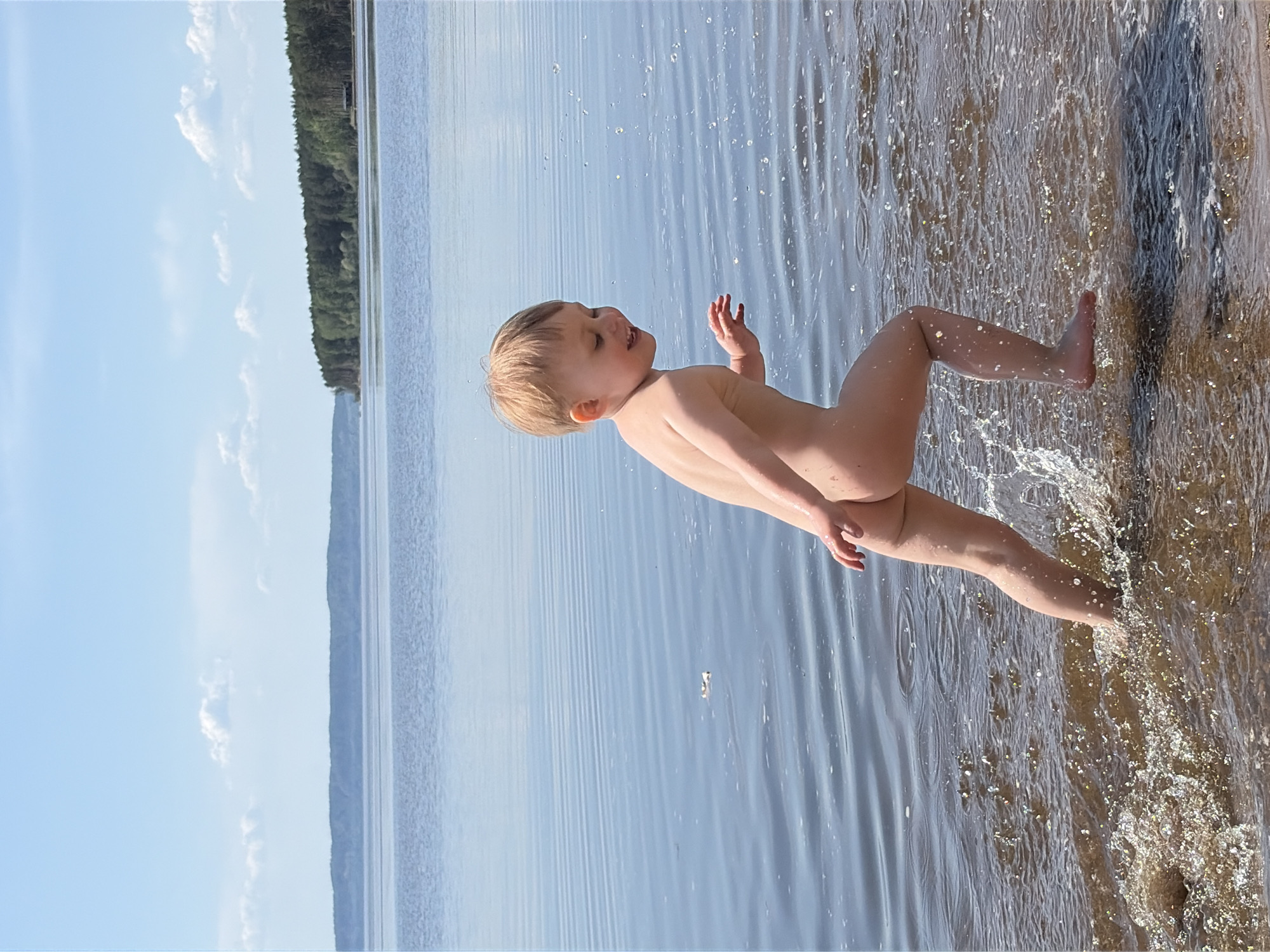 A young, nude child is standing in the shallow water along the shoreline of a lake. The child is fair-skinned with blonde hair and is facing left, with one arm raised and the other down. Splashes of water surround the child's legs as they appear to be moving. The lake is calm, with a slight ripple effect, and reflects the blue sky with a few clouds. A treeline is visible on the opposite shore. The location is Zabegaevo, Russia.
