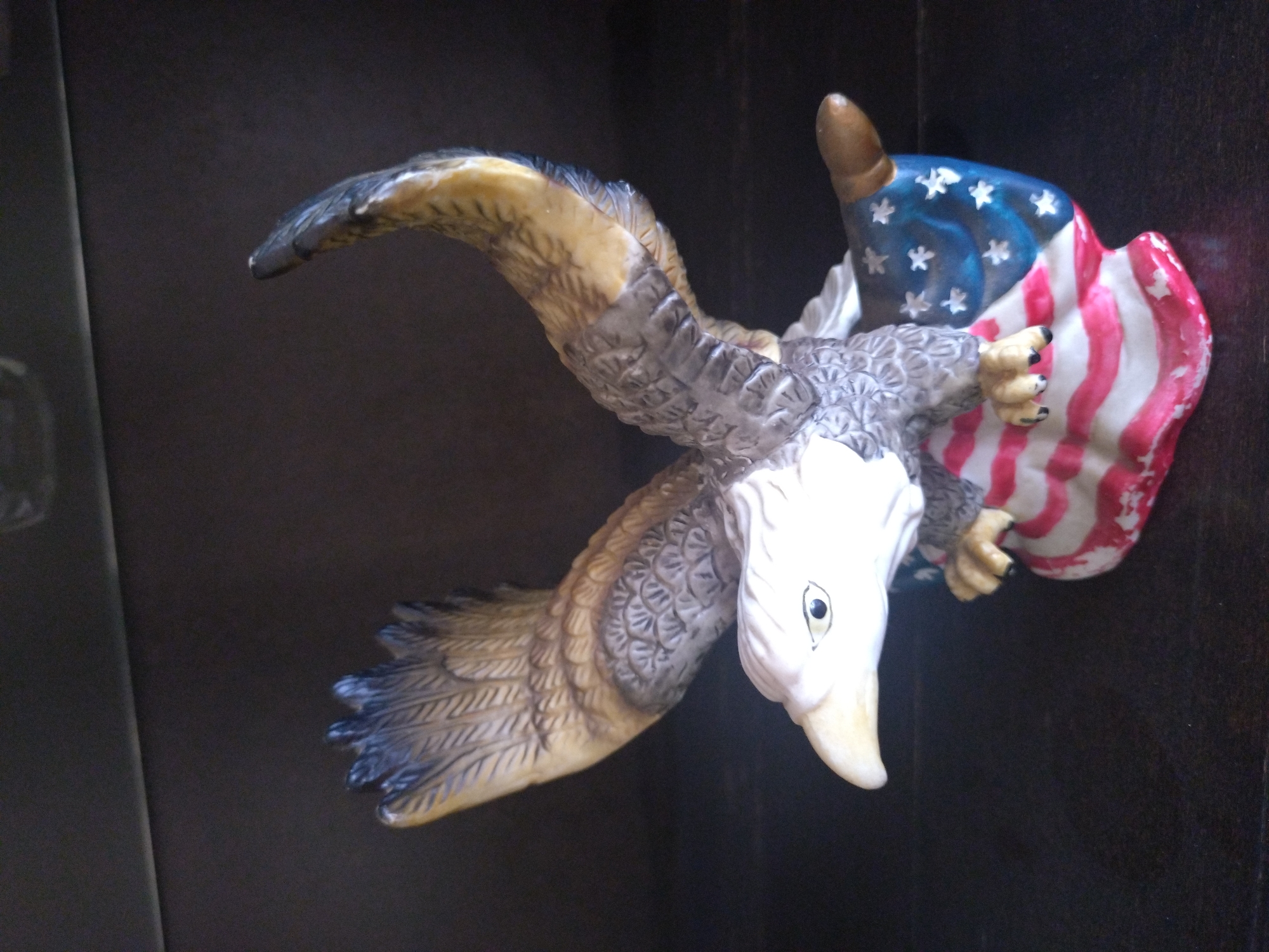 A ceramic figurine of an eagle is displayed on a dark surface. The eagle is depicted with its wings spread and is perched on an American flag. The figurine is detailed with feathers and has a white head with a yellow beak and black eyes. The flag shows red and white stripes and blue with white stars. The background is dark and out of focus, suggesting an indoor setting. There is no indication of a specific location in Stara Zagora, Bulgaria.