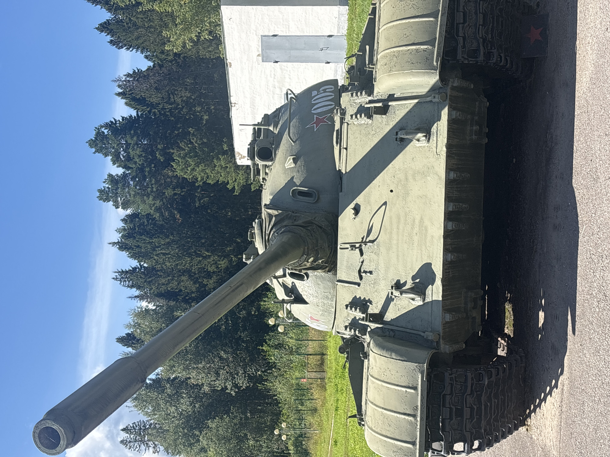 An olive-drab tank, identified by the number "005" and a red star marking, is parked outdoors on a paved surface in front of a white, rectangular building with a gray door. The tank is angled slightly towards the viewer, with its large barrel extending diagonally upwards and to the left. The tank's tracks are visible, and the surrounding environment includes a grassy area, a fence, and a dense line of tall evergreen trees under a clear blue sky with scattered white clouds. The scene appears to be a static display, possibly a museum or memorial site, in the city of Gamy, Russia.