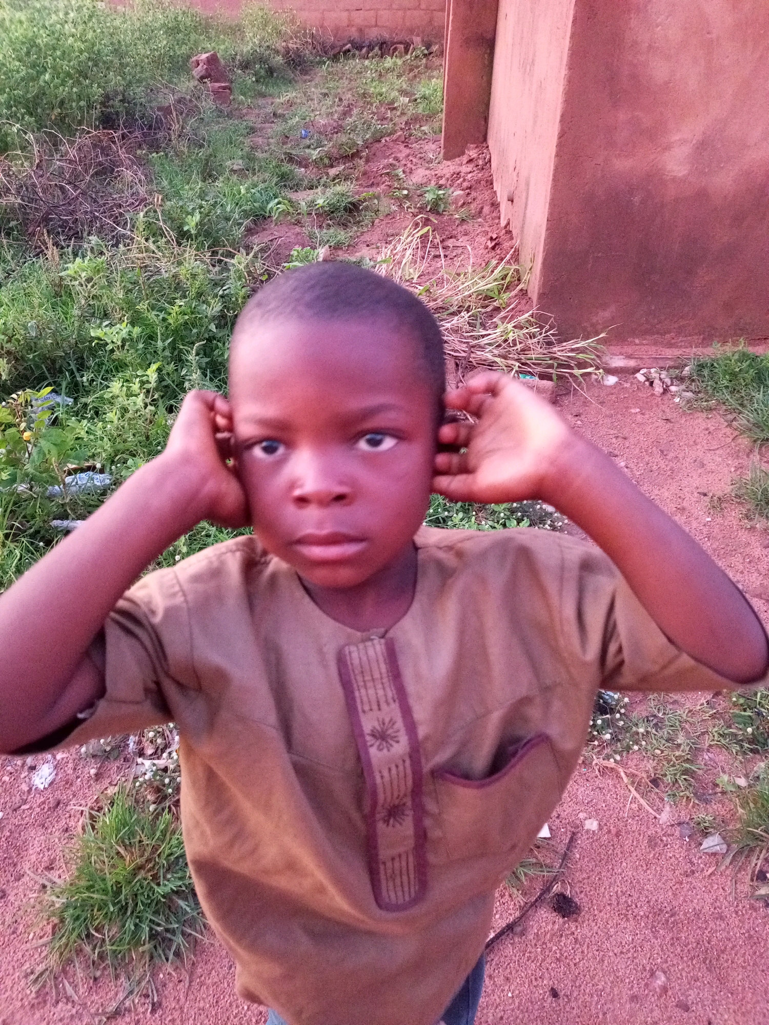 A young boy, appearing to be of African descent with dark skin and hair, stands outdoors in what is likely a rural or semi-urban area in Tempure, Nigeria. He is wearing a light brown, long-sleeved tunic with decorative maroon stitching and a small pocket on the left side of the chest. The boy is looking directly at the camera with a neutral expression. He has his right hand placed against his right ear and his left hand similarly placed against his left ear.
The environment surrounding the boy consists of dry, reddish-brown soil and sparse green vegetation, including grasses and some brush. In the background, a portion of a building constructed with rough, reddish-pink material, possibly mud brick or stucco, is visible on the right side. The ground is a mix of sand, small stones, and dry plant matter. The lighting suggests it may be late afternoon or early morning, with a slight reddish cast to the overall scene. The overall impression is of a candid portrait taken in a simple, outdoor setting.