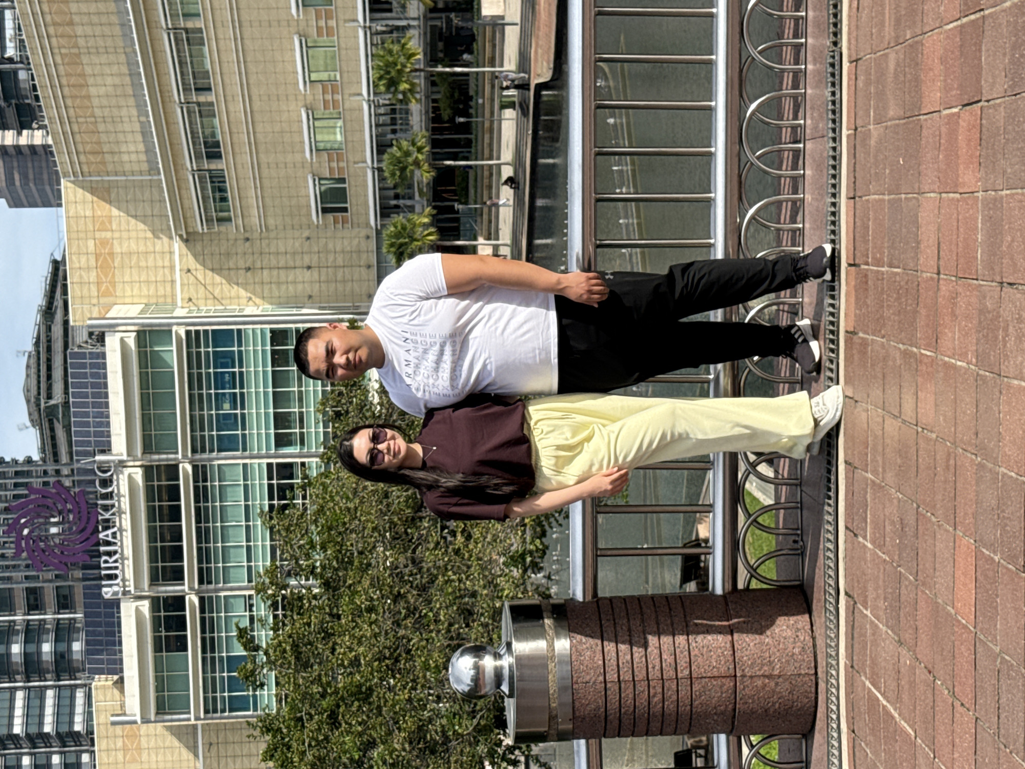 A photo of two people, likely a couple, standing on a brick-paved walkway in Kuala Lumpur, Malaysia. In the background is a building with a sign displaying "SURIA KLCC". The man is wearing a white t-shirt and black pants and is positioned slightly behind the woman. The woman is wearing a dark top and light yellow pants. A metal railing is visible in the foreground, separating the walkway from a body of water. The overall impression is a sunny day in a modern urban setting.