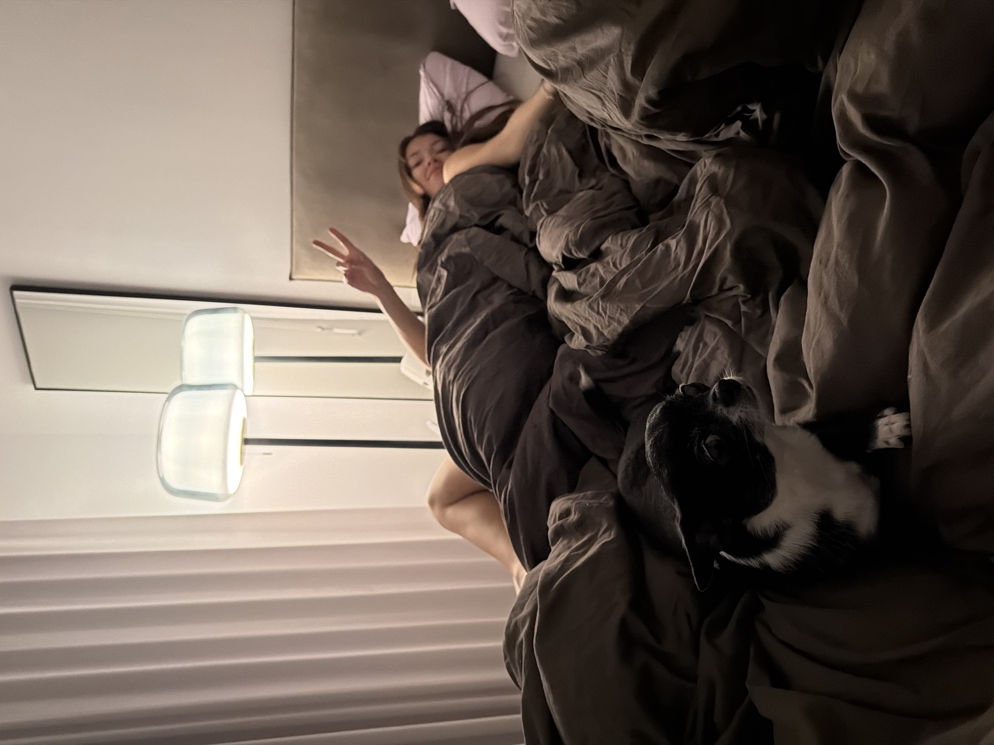 A woman lies in a bed with gray linens, giving a peace sign with her right hand. A black and white dog is lying next to her on the bed. A lamp is affixed to the wall near a white window with horizontal blinds. The location in the city of Tel-Aviv, Israel, cannot be determined from the image.