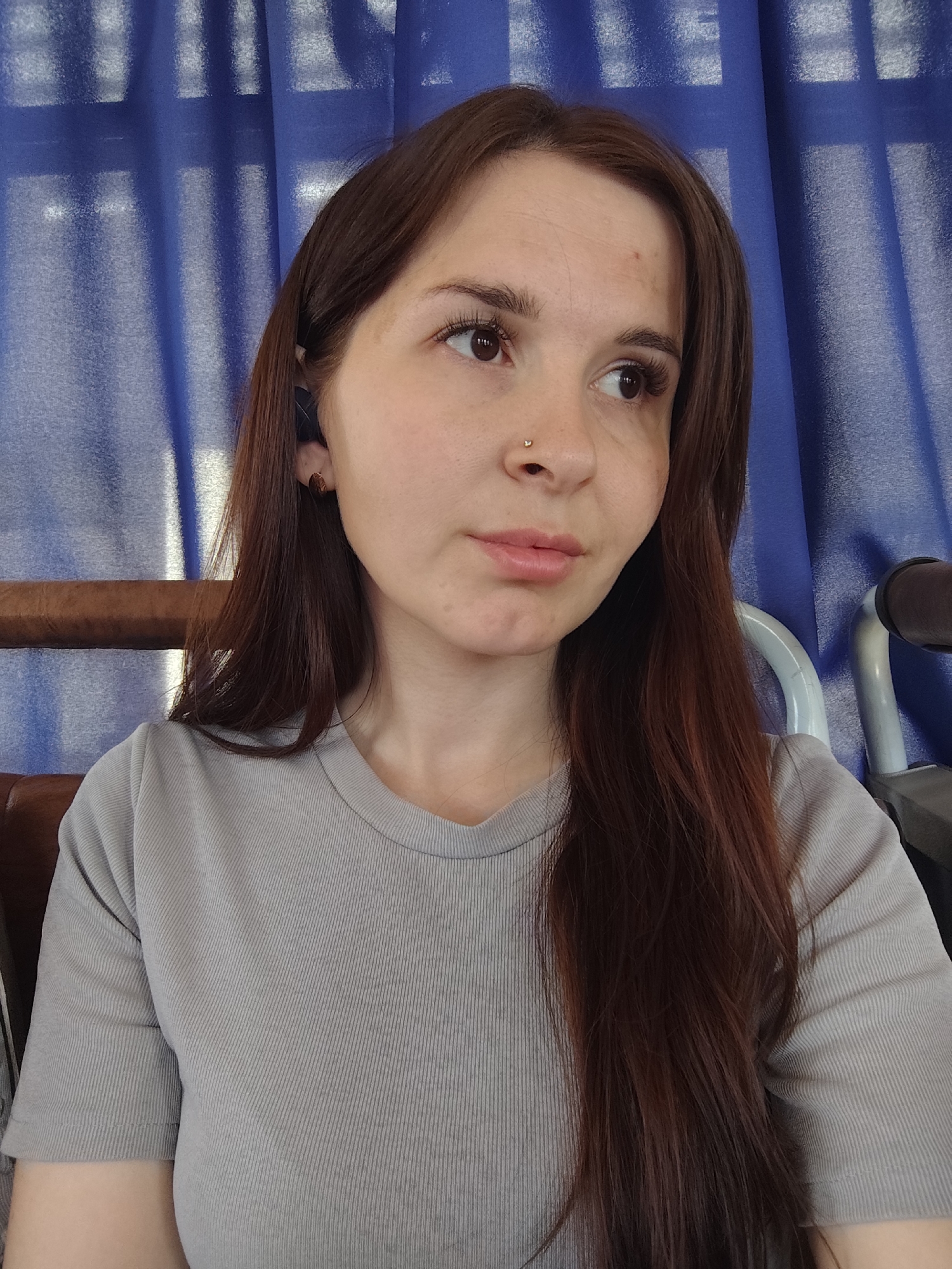 A woman is seated indoors with her head and upper torso visible. She has long, brown hair, and a nose piercing. She is wearing a grey ribbed t-shirt. Behind her are blue curtains. The location is Chaikovsky, Russia.