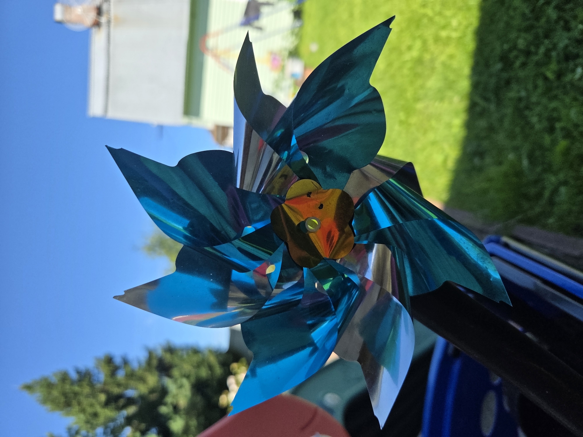 A metallic blue pinwheel is held in the foreground, its iridescent blades reflecting sunlight. The pinwheel is positioned against a backdrop of a clear blue sky, a green lawn, and residential buildings. In the distance, a tall green tree is visible on the left, and a house with a light-colored facade and a chimney is on the right. The overall setting suggests an outdoor environment, possibly a backyard in a residential area. No individuals are present in the image.