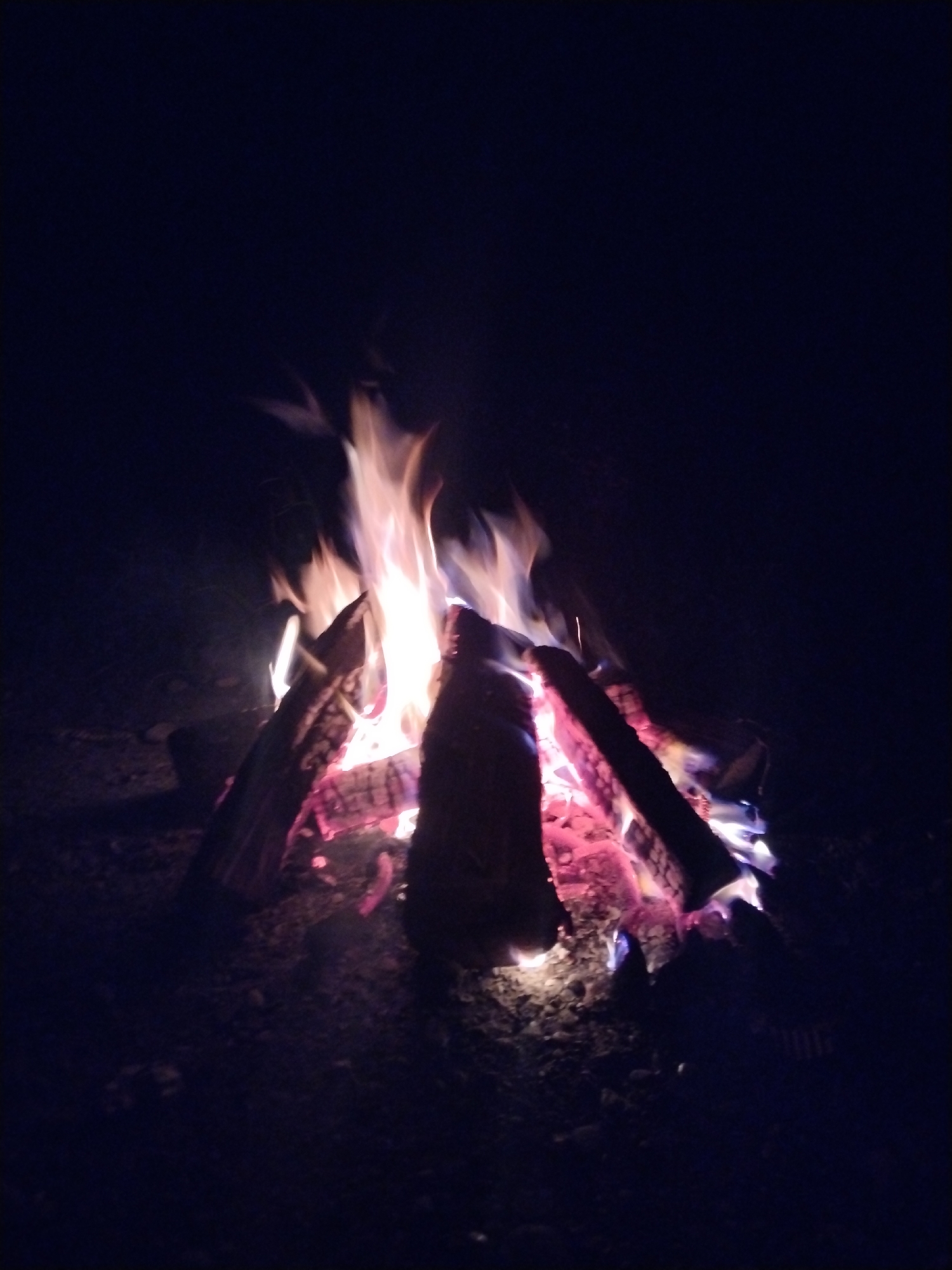 A campfire burns brightly in the darkness. Logs are stacked in a pile, with flames reaching upwards, casting a warm glow of red, orange, and blue onto the surrounding area. The ground beneath the fire appears to be dirt and gravel. The background is completely dark, suggesting the image was taken at night. The location of Бчара ақыҭа ахадара, Abkhazia, is not visible in the image.