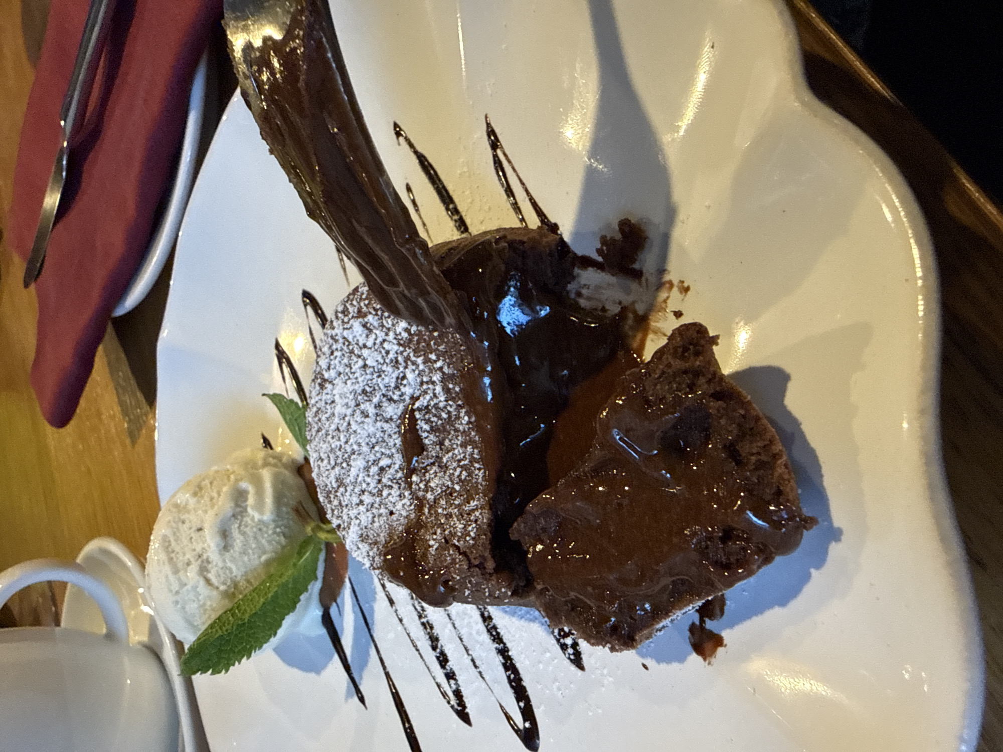 A chocolate lava cake is served on a white plate with vanilla ice cream and a sprig of mint. Chocolate sauce is drizzled on the cake and plate. The cake is dusted with powdered sugar. A fork and a red napkin are visible on a separate plate in the background. A white cup with a handle is also visible. The location is not discernible from the image.