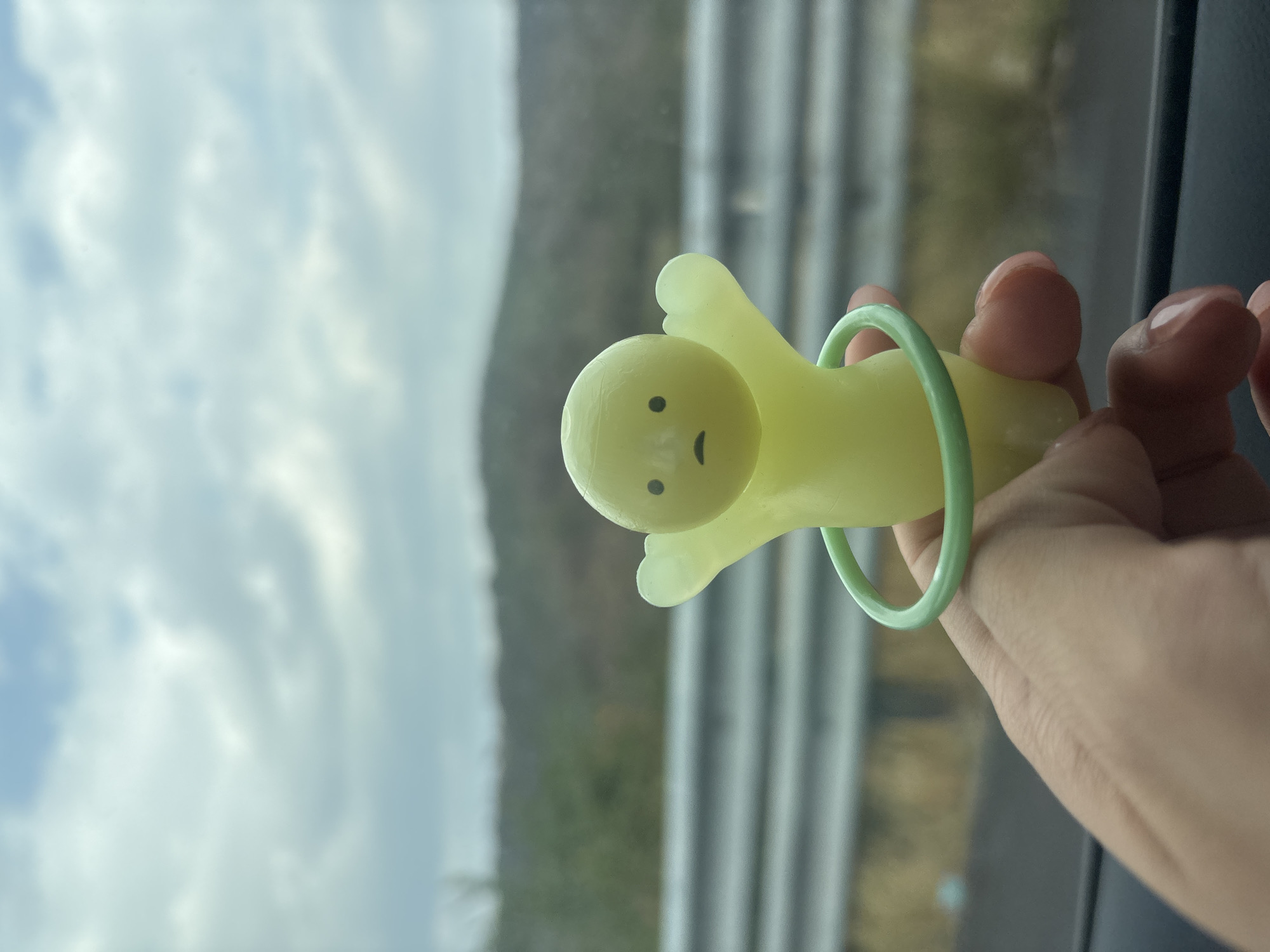 A hand holds a yellow, cartoonish, human-shaped toy with a green ring around it. The toy has two dots for eyes and a curved line for a mouth. The background shows a view from a window, with the sky and clouds visible. There is also a blurred view of what appears to be trees and/or vegetation outside the window. The city of San Felipe Cuapexco, Mexico is not discernible from the image.