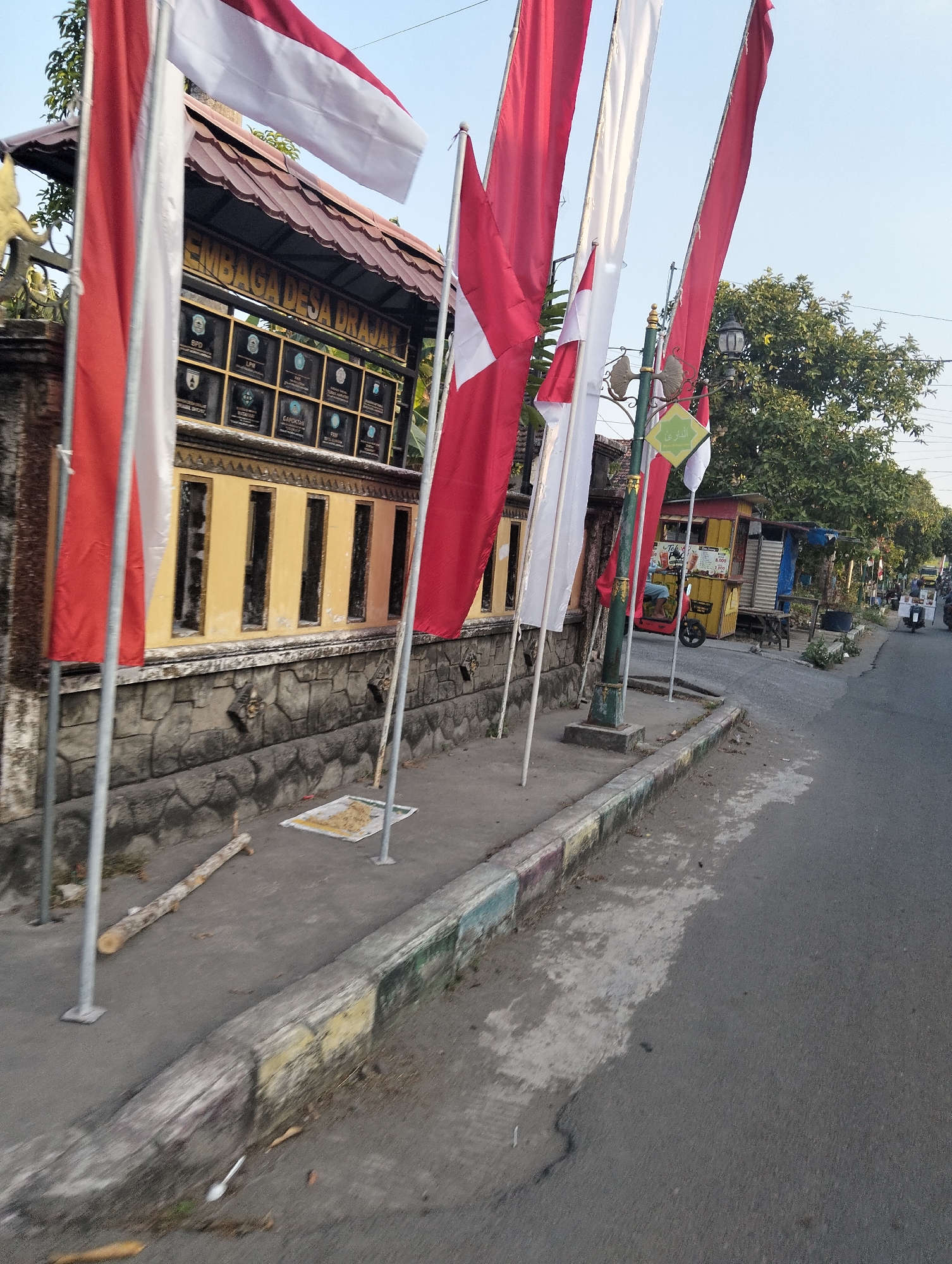 The image shows a street in Drajat, Indonesia, with several red and white flags displayed. The flags are on poles and appear to be related to a local government building, indicated by the sign "EMBAGA DESA DRAJAT" above a wall with several plaques. The building has a yellow facade with dark rectangular openings. Adjacent to the building is a sidewalk with a curb painted in various colors. A street runs alongside the sidewalk, with trees and a few small stalls or shops visible in the background. A small red vehicle, possibly a motorcycle with a sidecar, is parked near one of the stalls. There are no people clearly visible in the foreground, but a person can be seen inside the red vehicle. The overall scene depicts a street in a village or small town in Indonesia, with elements of local administration and commerce.