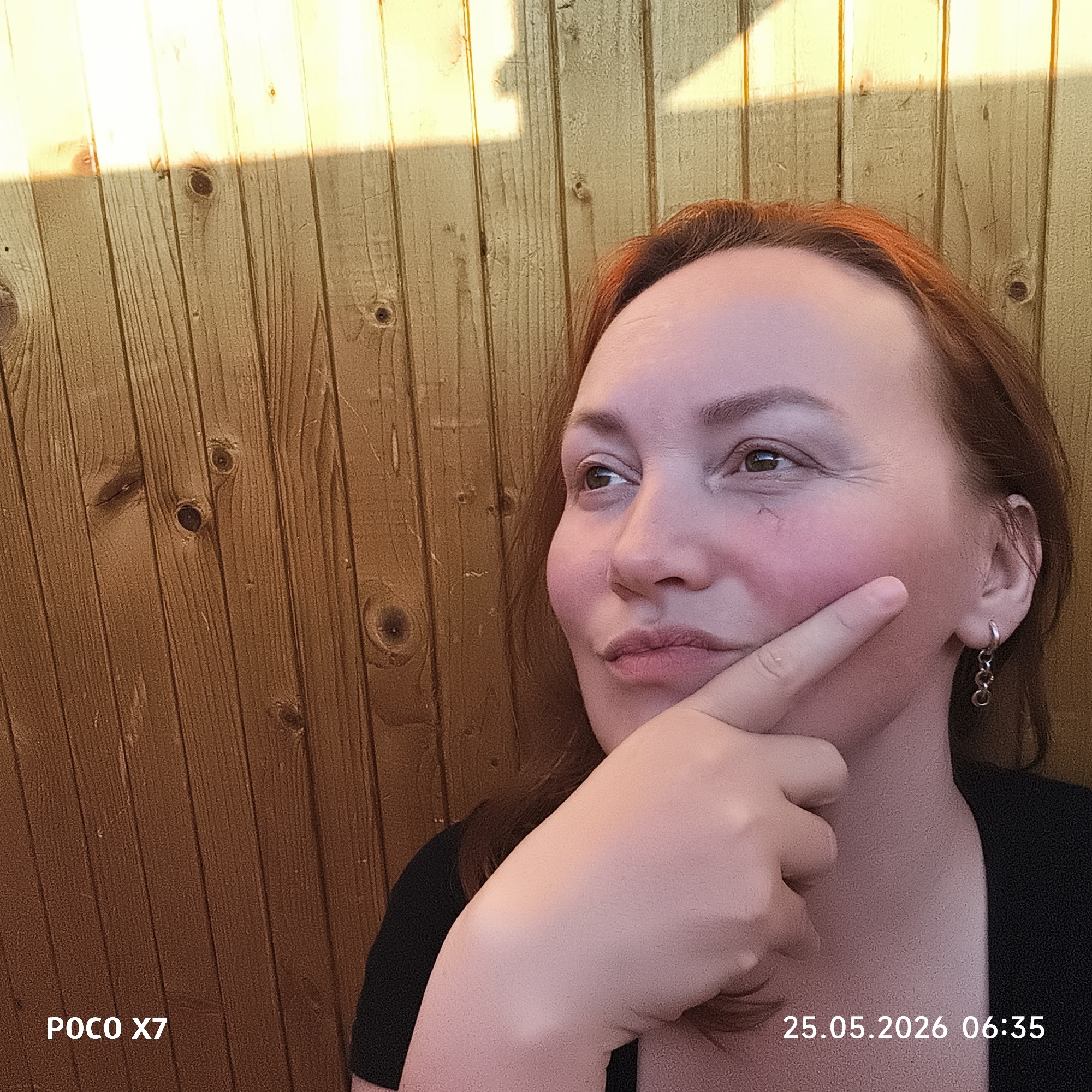 A woman with reddish-brown hair is shown from the chest up, in front of a wooden wall. She is looking off to the left with a thoughtful expression, her index finger touching her right cheek. She is wearing earrings and a black top. The background is a wooden wall, partially lit by sunlight. Text at the bottom indicates the image was taken on a "POCO X7" on "25.05.2026 06:35." The location is not specified, rendering the city as Unknown, Unknown.