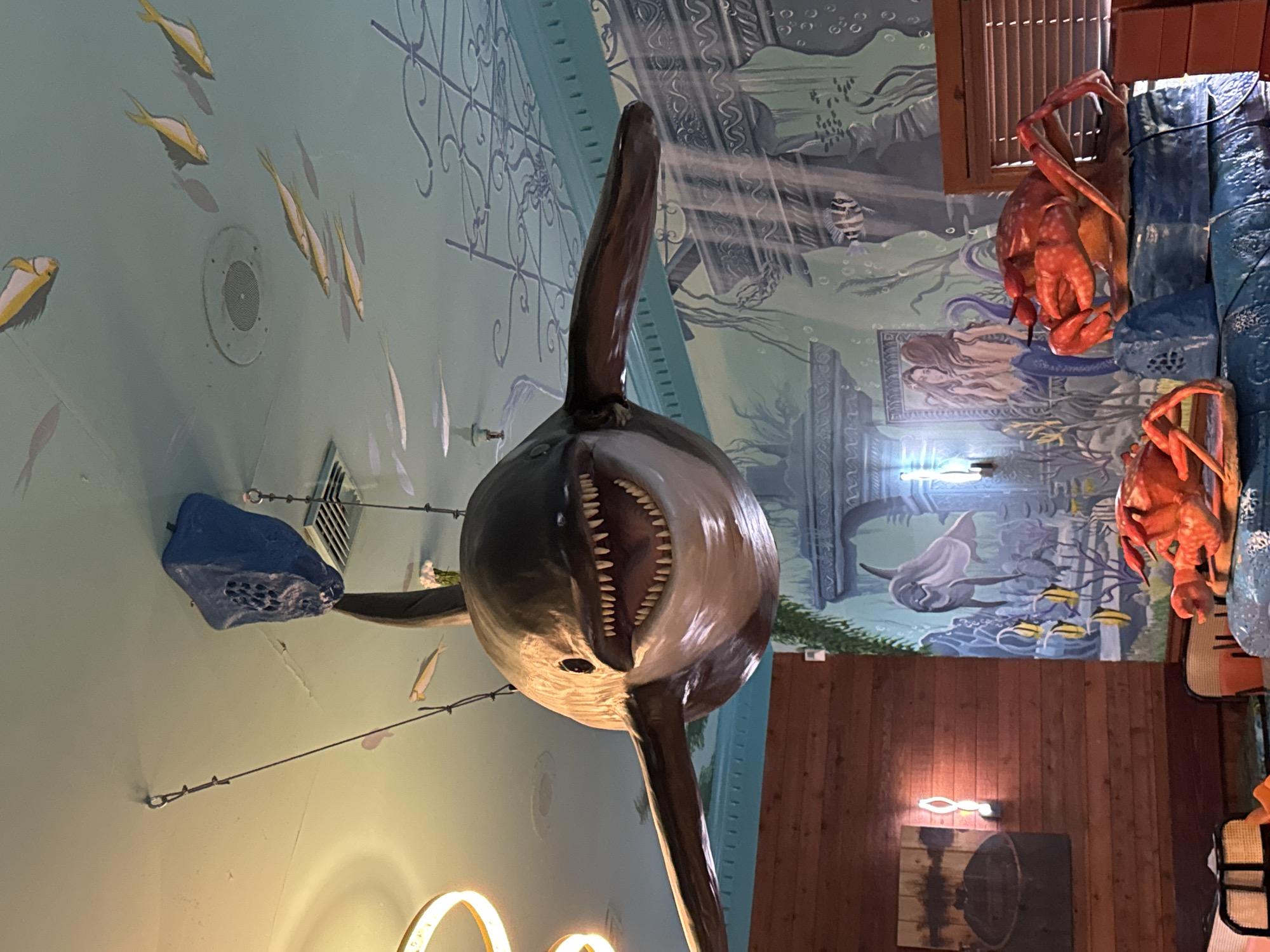 A wide-angle, eye-level shot depicts an indoor scene in Garden City Beach, USA, featuring an underwater-themed decor. A large, shark statue with its mouth open dominates the center, suspended from the ceiling. A blue speaker is visible to the left. A wall mural with undersea imagery and crab statues is on the right. The right wall is also paneled in wood and has framed art.
