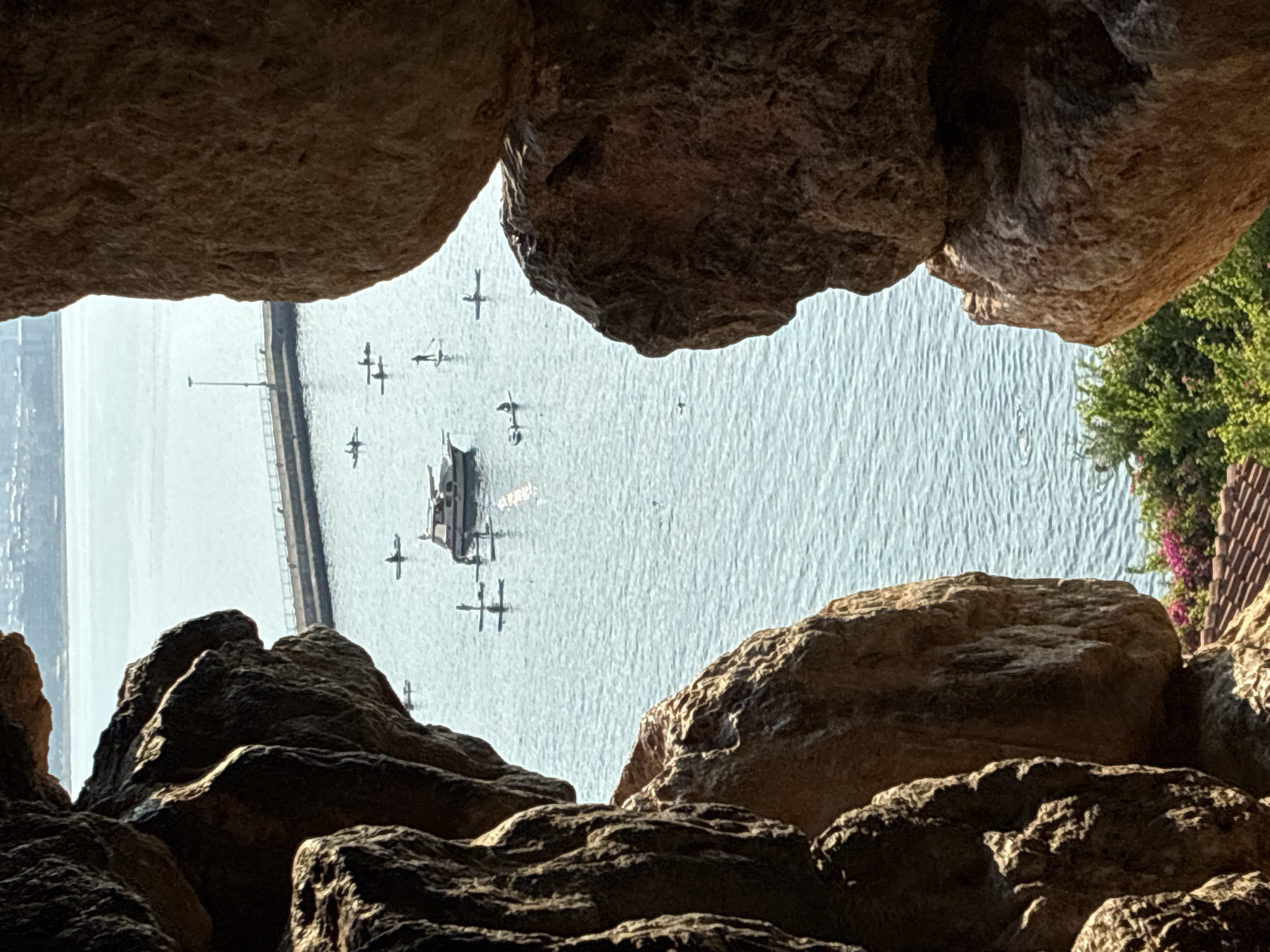 The image captures a view of the Alanya harbor in Alanya, Turkey, framed by large rock formations. In the foreground, rough, textured rocks create a natural frame, with one on the left and two on the right. Beyond the rocks, the clear blue water of the harbor is visible. Scattered across the water are several individuals on paddleboards, some standing and others seated. A small motorboat is anchored in the middle of the harbor, with people on board or near it. In the distance, a pier extends into the water, with a lamppost at its end. The background shows the hazy outline of buildings and hills, suggesting the presence of the city. Lush green trees and vibrant pink flowers are visible in the lower right portion of the image, with terracotta roof tiles hinting at structures below. The scene suggests a sunny day with moderate activity in the harbor.