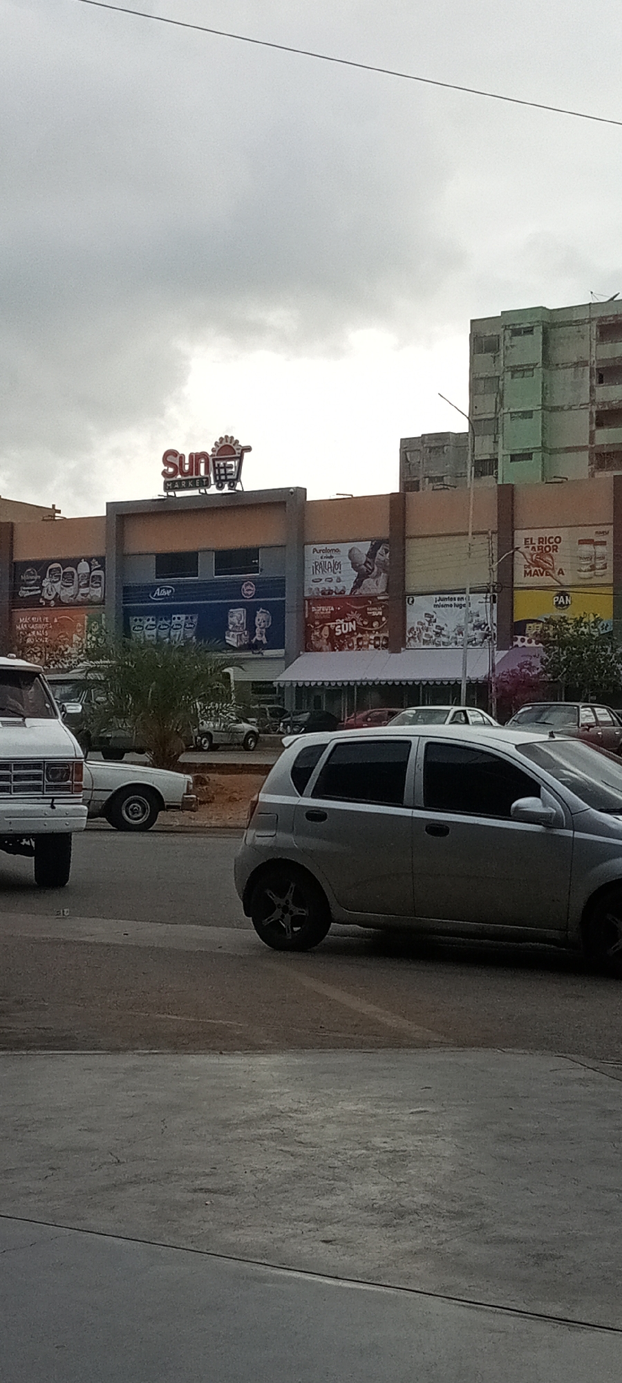 The image presents an overcast urban scene, dominated by a multi-story commercial building that features the "Sun Market" logo prominently displayed on its upper facade. The building's exterior walls are adorned with various advertisements and promotional posters.
A street runs in front of the building, and several vehicles are visible, including a white van and a silver hatchback car in the foreground. Other vehicles are visible in the distance, parked near the market.
The backdrop includes a tall, older apartment building with a green facade, indicative of an established urban environment. The overall composition suggests a scene captured on a city street near a commercial center.