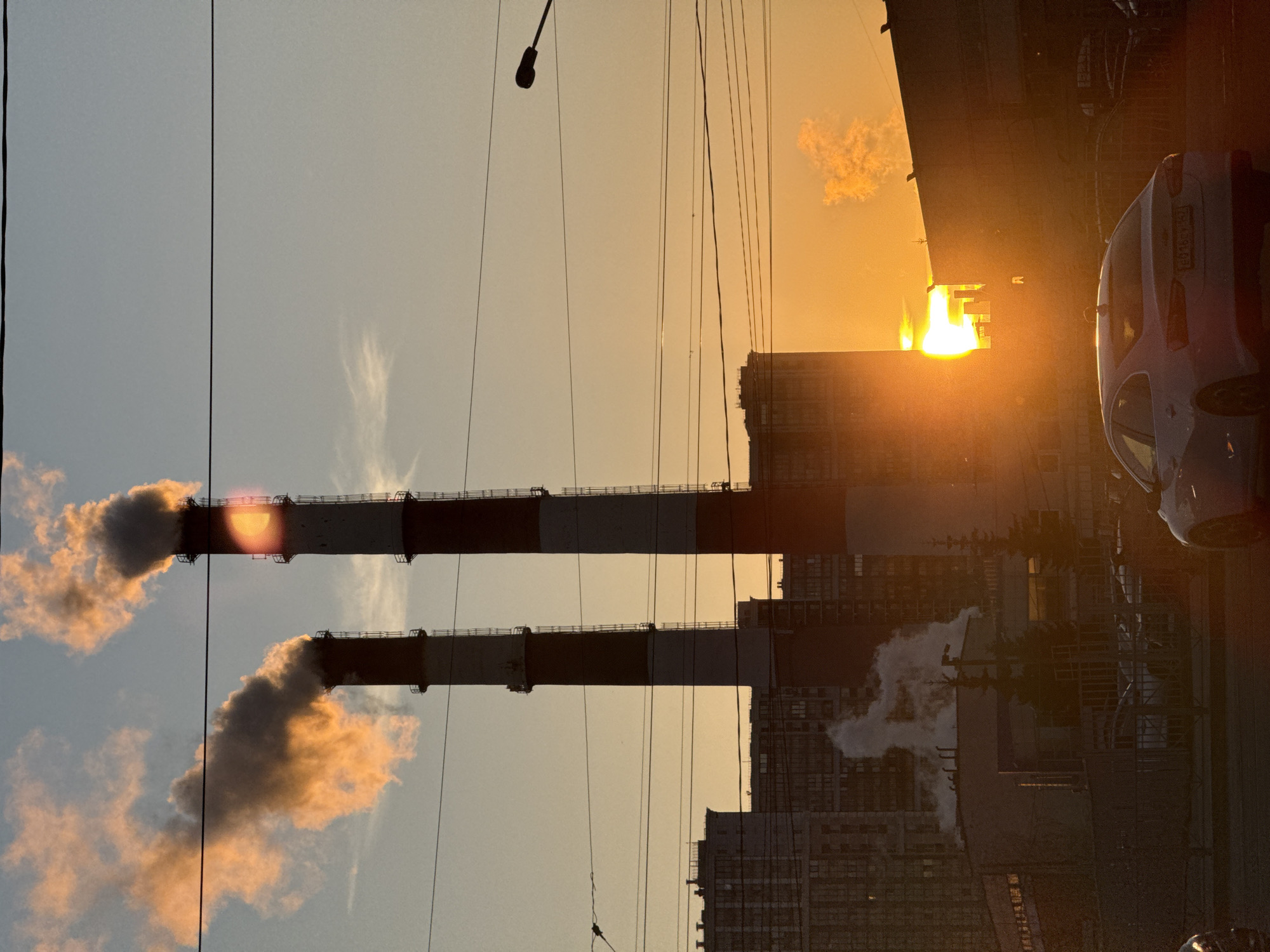 The image shows an industrial area with two large smokestacks emitting plumes of smoke. The smokestacks are painted with alternating red and white bands. In the background, there are buildings that appear to be part of a city. The scene is backlit by a bright setting or rising sun, casting a warm glow. There is also a car parked on the street to the right of the image. Given the details in the image and the prevalence of such industrial zones in St. Petersburg, Russia, the location is likely within an industrial district of the city.