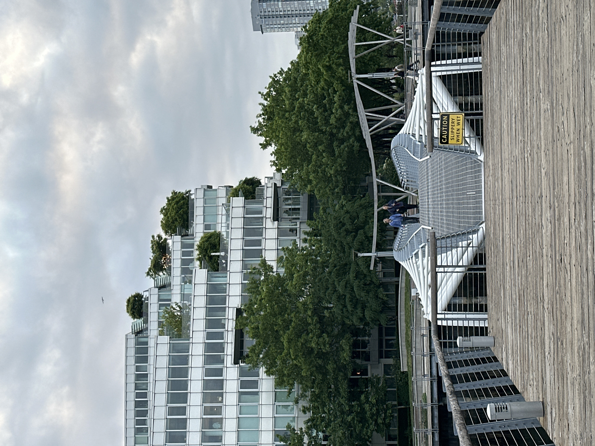 The image depicts a modern architectural scene, possibly in Vancouver, Canada, given the distinctive building style with rooftop gardens. The foreground shows a wooden boardwalk with a metal grate staircase and an artistic, spiraling white structure. A yellow "CAUTION SLIPPERY WHEN WET" sign is visible on the structure. Two people are walking on the walkway. To the left, a tall building featuring many windows and stepped terraces is seen, some terraces adorned with greenery. In the background, a taller building is also visible. The sky is overcast, suggesting a cloudy day. A bird is flying near the edge of the building.