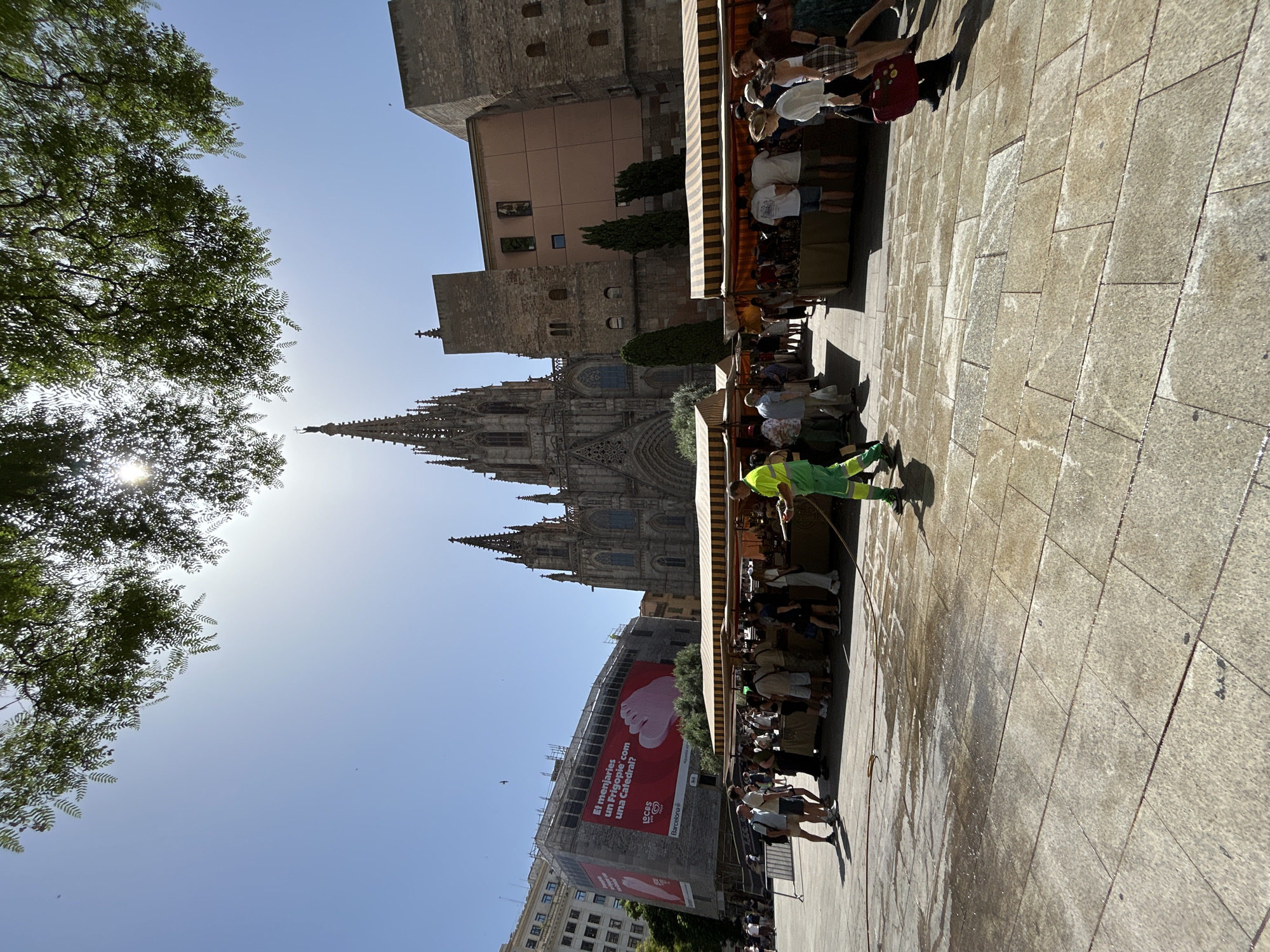 The image depicts an outdoor scene in Barcelona, Spain, under a bright, clear sky, suggesting it is daytime and likely warm. In the background, the impressive Gothic facade of the Barcelona Cathedral is visible, with its intricate spires and architecture dominating the skyline. Adjacent to the cathedral, there is a tall, modern building covered in scaffolding and adorned with a large red billboard.
In the foreground, a paved plaza is the main setting. A person wearing a bright green vest is hosing down the stone tiles, creating wet patches on the ground. A line of people are gathered under awnings, possibly at market stalls or a cafe, observing the surroundings or interacting with each other. The composition is vertical, with a large tree on the left framing the view of the sky and the upper parts of the buildings. The sunlight streams through the tree's foliage, creating a lens flare effect.
The billboard on the building shows a close-up of a large, pink foot and text in Spanish. Parts of the text are legible: "El menjaries un frigopice 'com una Catedral?'" and a logo with "LOCUS" and "Barcelona".
The overall atmosphere appears to be bustling, with people enjoying an outdoor urban environment, with the historical cathedral providing a dramatic backdrop. The activity of the person cleaning the plaza suggests routine maintenance amidst the public gathering.