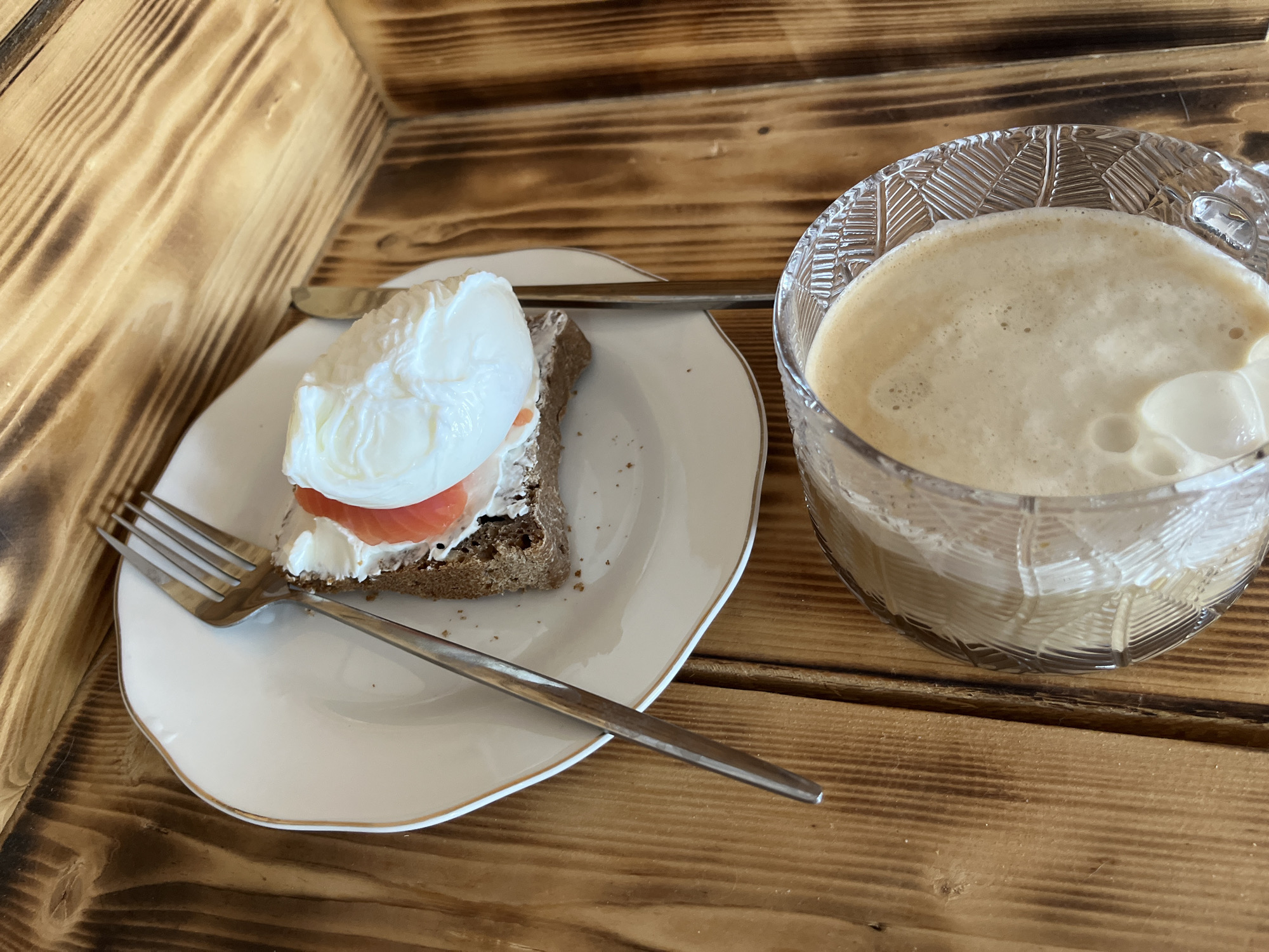 A close-up shot captures a rustic breakfast or brunch setting in Chaikovsky, Russia, featuring an open-faced sandwich and a beverage. The meal is presented on a textured wooden surface, possibly a table or tray, characterized by its prominent grain patterns and warm tones.
On a white ceramic plate with a subtle gold rim, an open-faced sandwich is prepared. It consists of a slice of dark bread, generously spread with a white, creamy cheese or spread, topped with a thin, folded slice of pinkish-orange salmon. A perfectly poached egg, with its bright white exterior and visible, gently bulging yolk underneath, crowns the sandwich. A silver-colored fork rests in front of the sandwich on the plate, while a matching knife is positioned behind it.
To the right of the plate, a clear glass mug, featuring an intricate, textured or cut-glass pattern, holds a light brown beverage with a substantial layer of frothy foam on top, indicative of coffee, a latte, or cappuccino. The foam has small bubbles and a smooth consistency, with a hint of a lighter, creamy element visible near the rim.
The scene suggests a cozy and inviting meal, likely consumed in the morning or early afternoon. There is no visible text in the image.