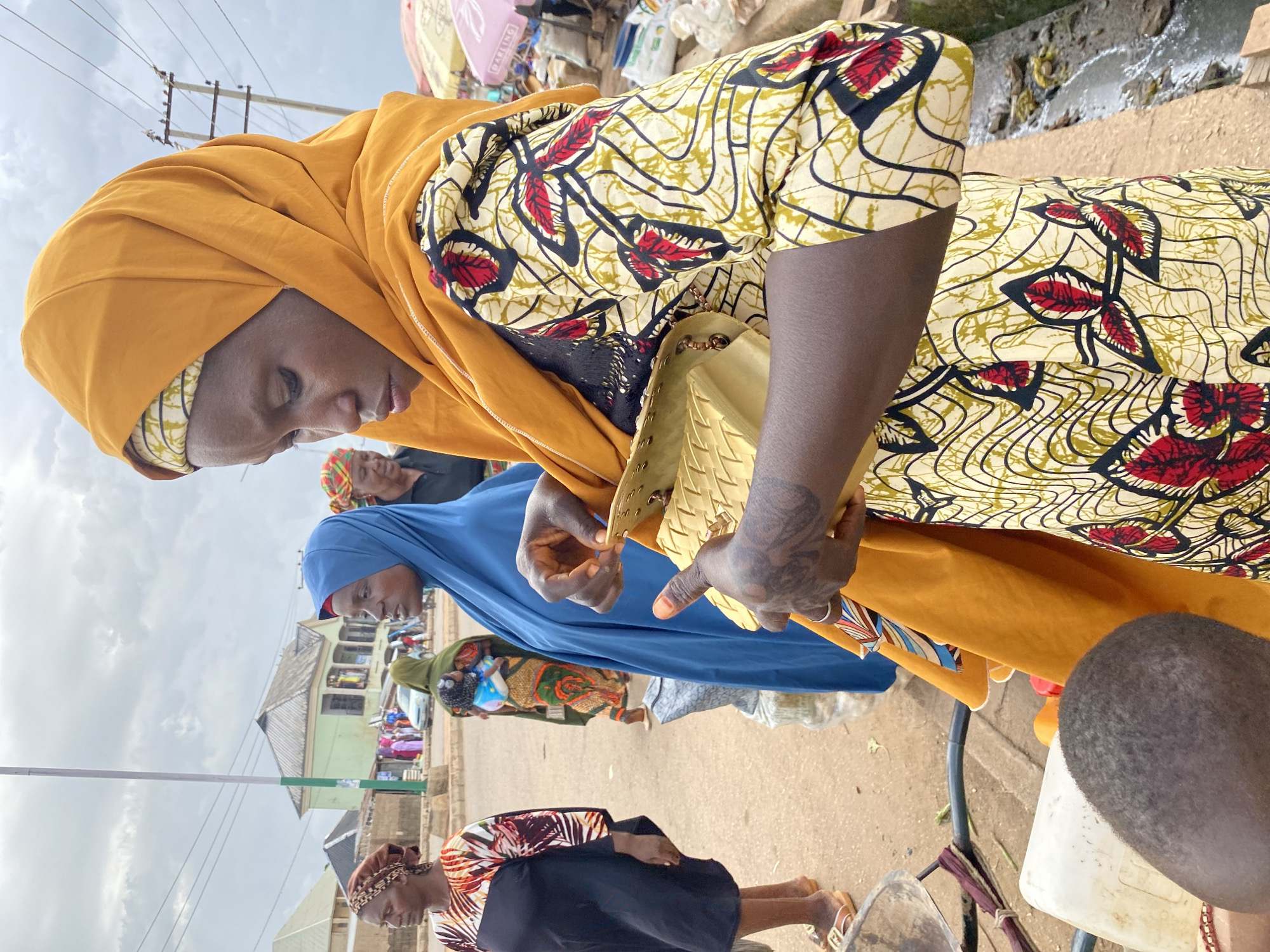 The media shows a close-up, side-view of a woman wearing a vibrant yellow headscarf and a patterned yellow, red, and black dress. She is holding a gold-colored, woven handbag. Her right hand, adorned with intricate tattoos and a ring, is in the process of closing the bag. She appears to be looking down, her expression unreadable.
Behind her, other individuals are partially visible, also dressed in colorful attire. One person is wearing a bright blue hijab, and another is in a green headscarf. The background suggests an outdoor market scene in Bukuru, Nigeria. There are hints of stalls with goods, electrical poles, and buildings. The sky is overcast, indicating a cloudy day. The overall impression is of a busy market environment with women engaged in shopping or selling. The time of day is likely daytime, given the lighting and activity. No specific text is clearly legible in the image.