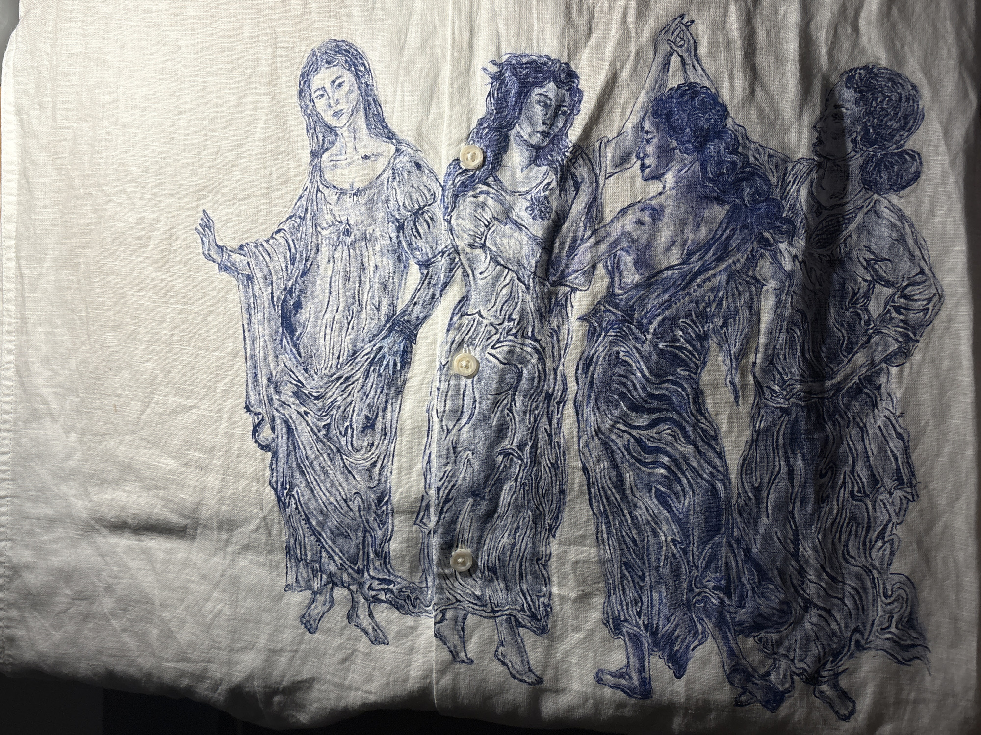 The image features a blue-ink drawing or print on an off-white, textured fabric, likely a piece of clothing such as a shirt, due to the presence of visible buttons. The artwork depicts five female figures arranged in a procession or dance, rendered in a classical or Renaissance-inspired style.
From left to right, the first figure stands facing forward with her left arm gently extended. The second figure stands beside her, also facing forward, with her arms seemingly held to her chest. Two small, round, light-colored buttons are sewn onto the fabric over her torso. The third and fourth figures are engaged in what appears to be a dance or graceful interaction, with their arms raised and possibly clasped together. The third figure faces right, while the fourth faces left, looking towards the center. The fifth figure, on the far right, faces generally right but looks back over her right shoulder.
All five women are depicted with long, flowing hair and are dressed in loose, draped garments reminiscent of classical antiquity. Their bare feet are visible below their long dresses, suggesting movement. The drawing style utilizes detailed lines and cross-hatching to create shading and texture. A third button is visible on the fabric below the second figure. The fabric itself shows subtle creasing and a visible woven texture. No text is visible in the image. The overall scene conveys a sense of elegant motion and timeless beauty.
