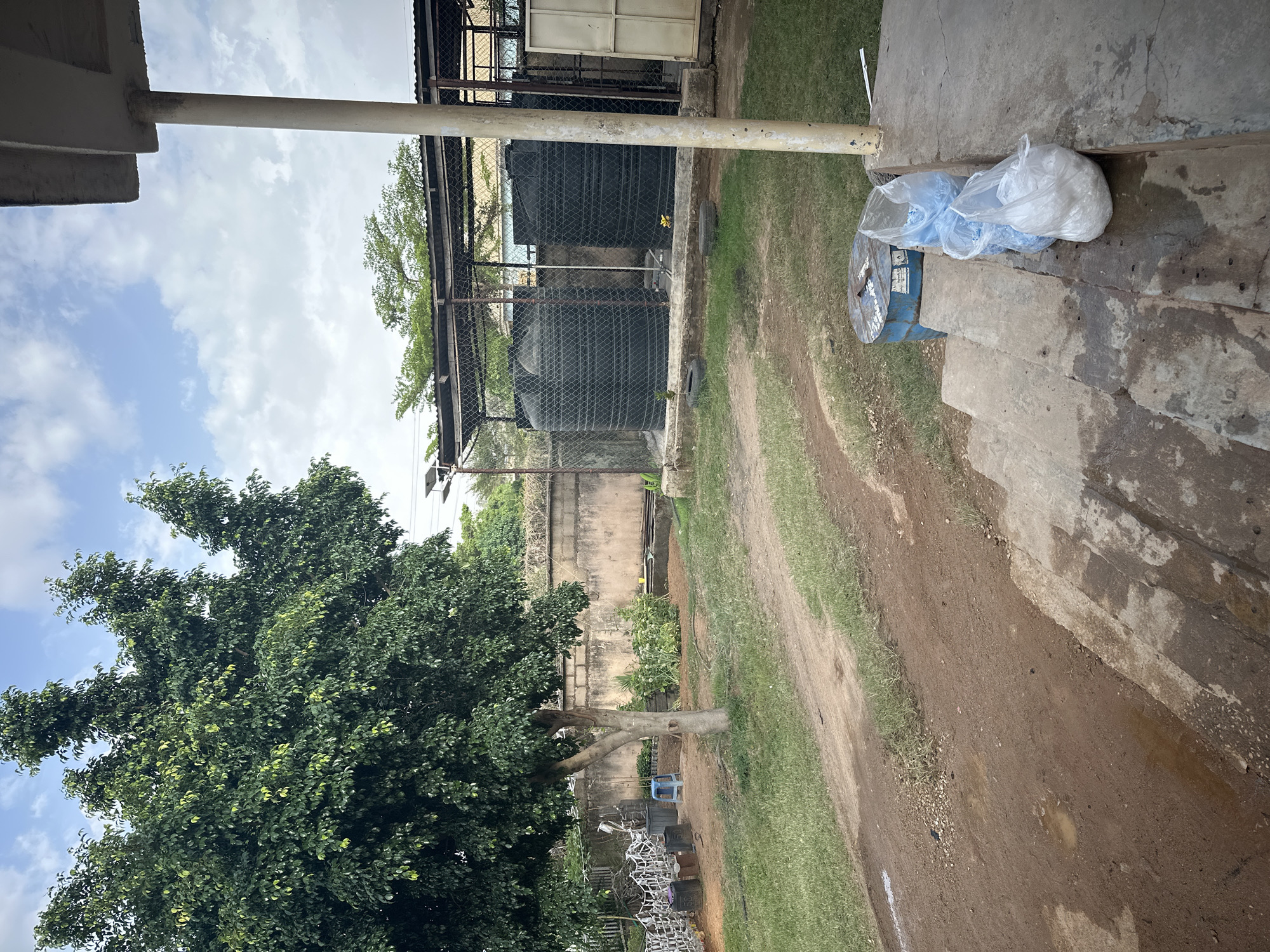 This image shows a scene in Jos, Nigeria, likely in a semi-urban or industrial area. In the foreground, there is a concrete structure with steps, upon which lie several plastic bags filled with material, possibly rice or grain. A blue container or drum sits adjacent to the bags. To the left, a large, mature tree with dense foliage dominates the view. Behind the tree, there is a fence and a glimpse of what appears to be stacked metal objects. Further back, large water storage tanks are enclosed by a chain-link fence. A dirt road or path cuts through the grassy area. The sky is partly cloudy. No people are visible.