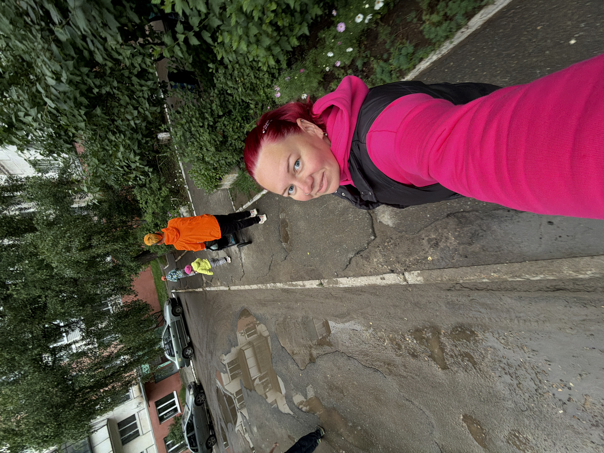 A woman with bright red hair, wearing a bright pink hooded sweatshirt and a black vest, is taking a selfie while walking outdoors. She is looking at the camera with a slight smile. In the background, a woman in an orange hooded jacket is pushing a stroller, and a small child in a yellow coat is walking beside her. There are two cars parked on the left side of the image, and the ground is wet with puddles, suggesting recent rain. The scene appears to be a residential area with trees and a building in the background. The image was taken on Lenina Street in Chaikovsky, Russia.