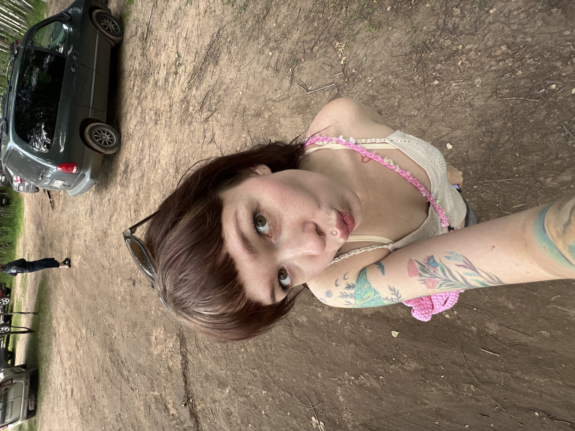 A woman with dark brown hair and bangs, wearing a cream-colored tank top with pink and green crochet straps, is taking a selfie outdoors. She has a lip piercing and several tattoos on her left arm, including a stylized blue bird and pink flowers. She is standing on a dirt path in a wooded area, with several vehicles visible in the background. A dark green minivan is parked to her right, and a silver van is parked in the distance to her left. A person in a dark jacket and pants is walking away from the camera in the background, near the silver van. The scene appears to be a park or a natural area, possibly near a campsite or parking lot. The presence of multiple vehicles suggests it could be a recreational area where people have driven to.