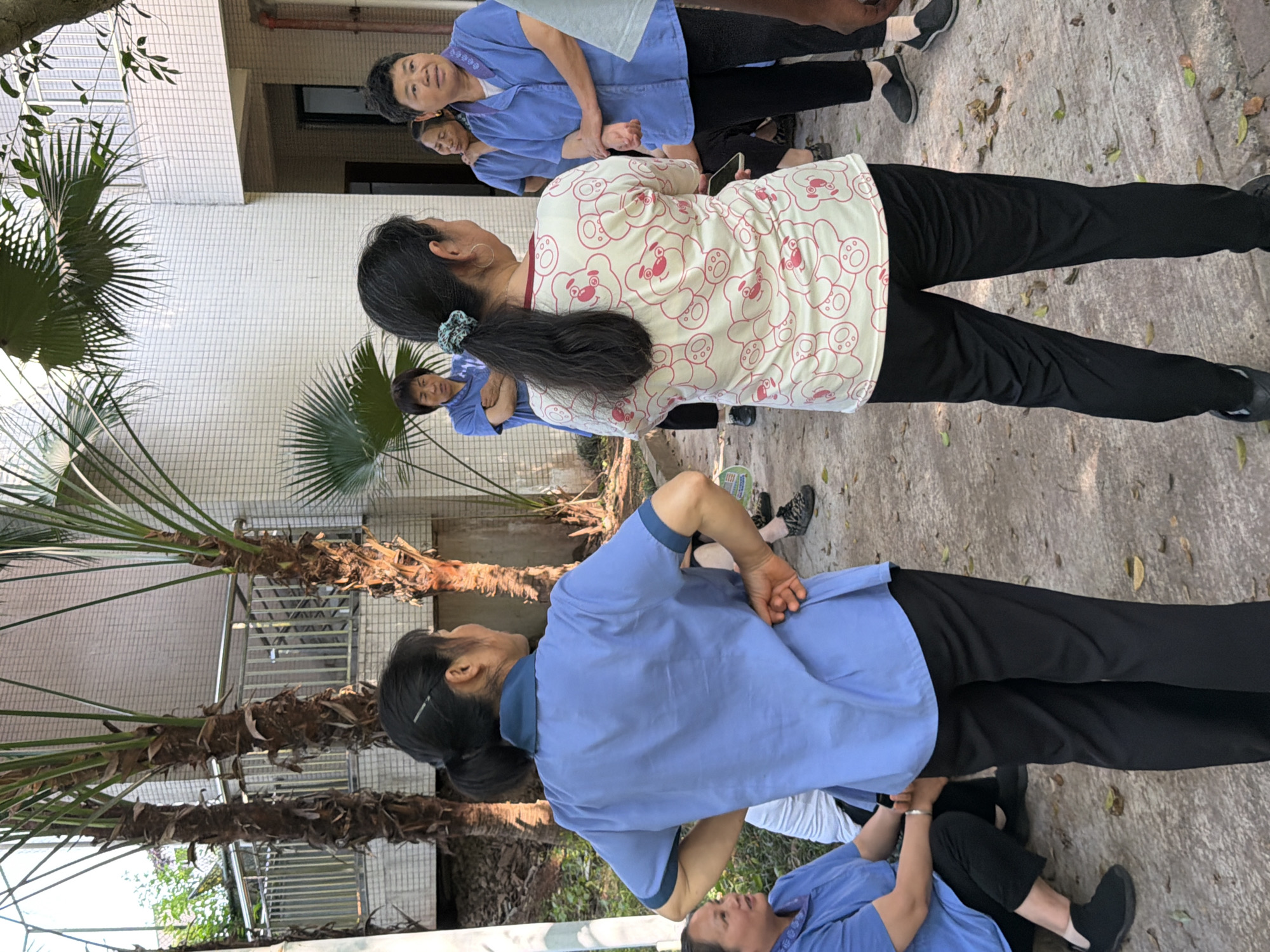 A group of women in blue uniforms and black pants are standing and stretching in an outdoor area. One woman is wearing a white shirt with red bears on it. They are near a building with white tiled walls and palm trees. The location is potentially in the city of Jiangjin, China.
