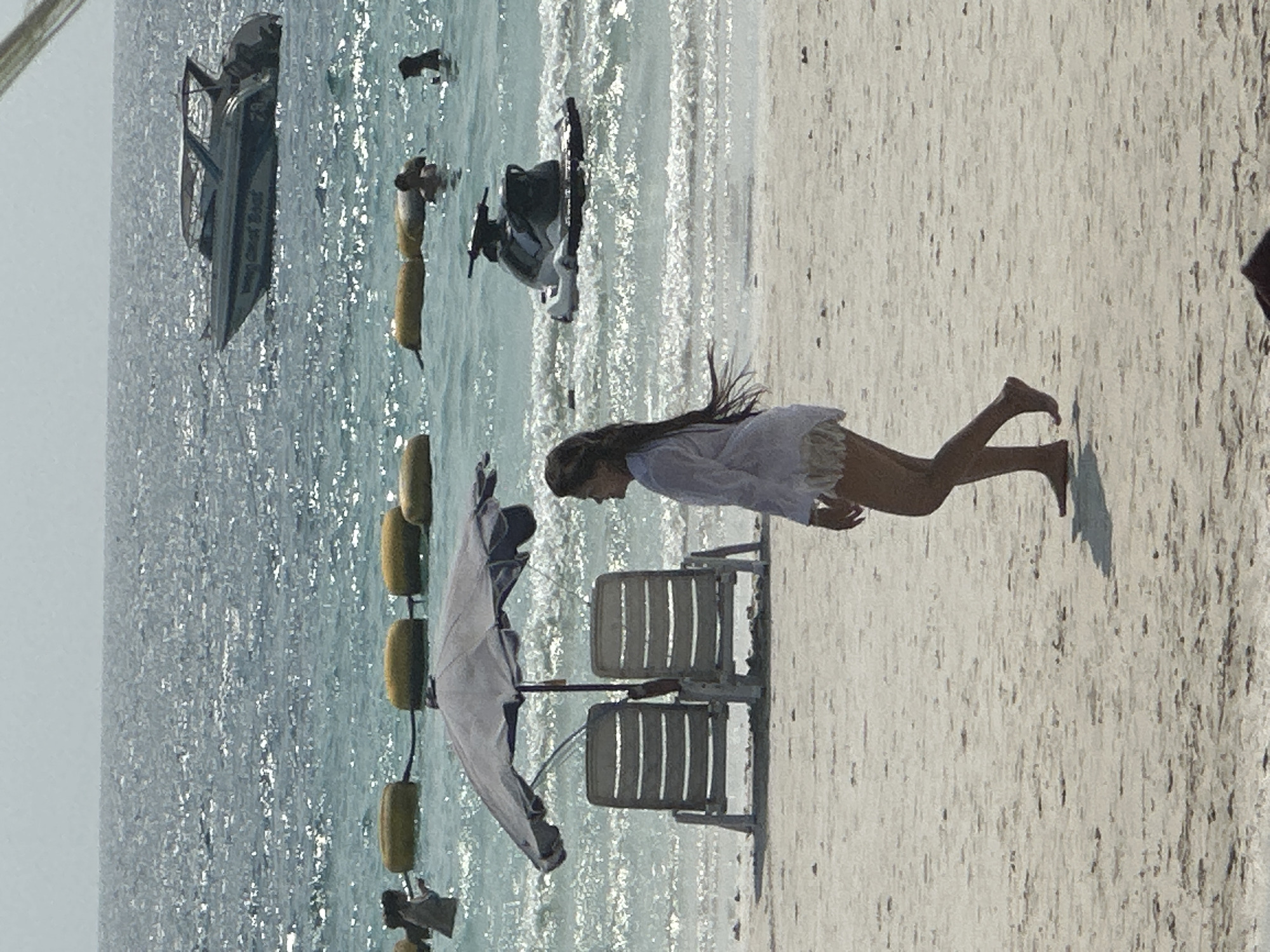 The image presents an outdoor scene at a beach in Pattaya City, Thailand. A woman is lying on the sandy beach near two beach chairs. Further into the water, a speedboat and a jet ski are visible. There are also yellow buoys in the water. Other figures are present in the water further away.