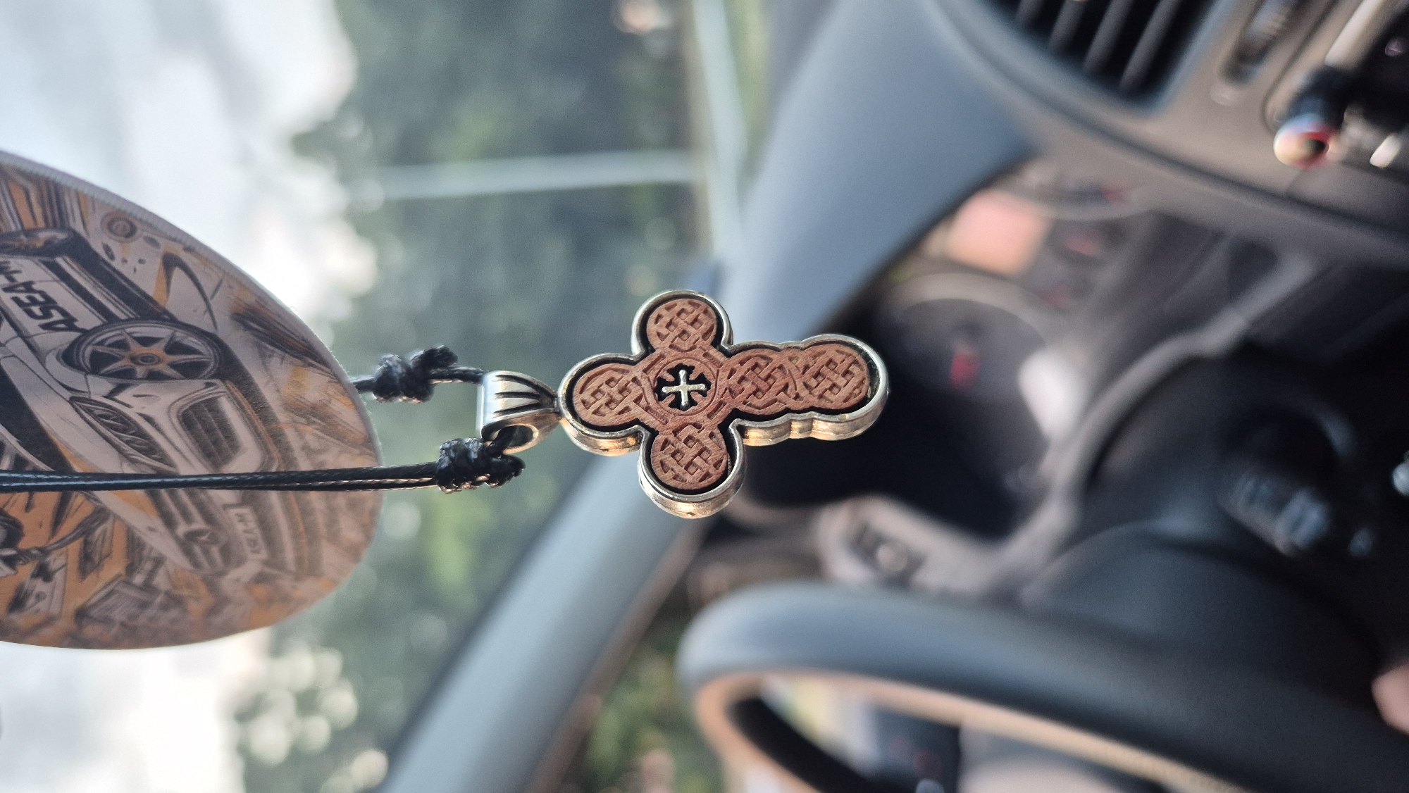 A close-up, shallow depth-of-field shot shows a decorative wooden and metal Christian cross hanging by a black cord inside a vehicle. The cross has intricate knotwork designs and a smaller cross in the center. Behind the cross, the interior of the car is visible, including the dashboard, steering wheel, and air vents. A circular car air freshener with an image of a sports car is hanging above the cross. The background outside the car is blurred, showing greenery and a bright sky. The image appears to be taken in Lovech, Bulgaria.